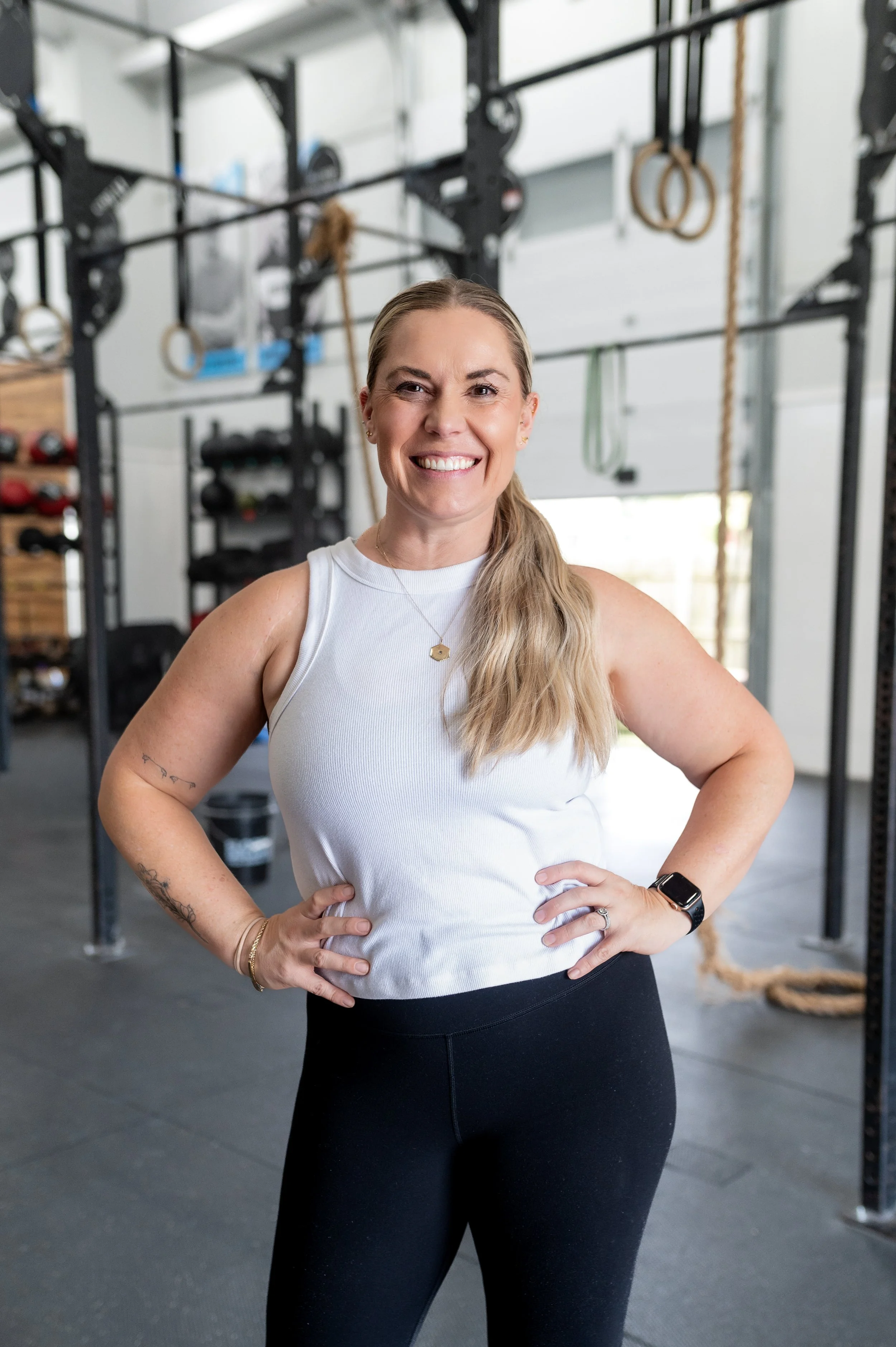 A woman with blonde hair tied back, smiling confidently in a gym, wearing a white tank top and black leggings, with gym equipment, including gymnastic rings and ropes, in the background.