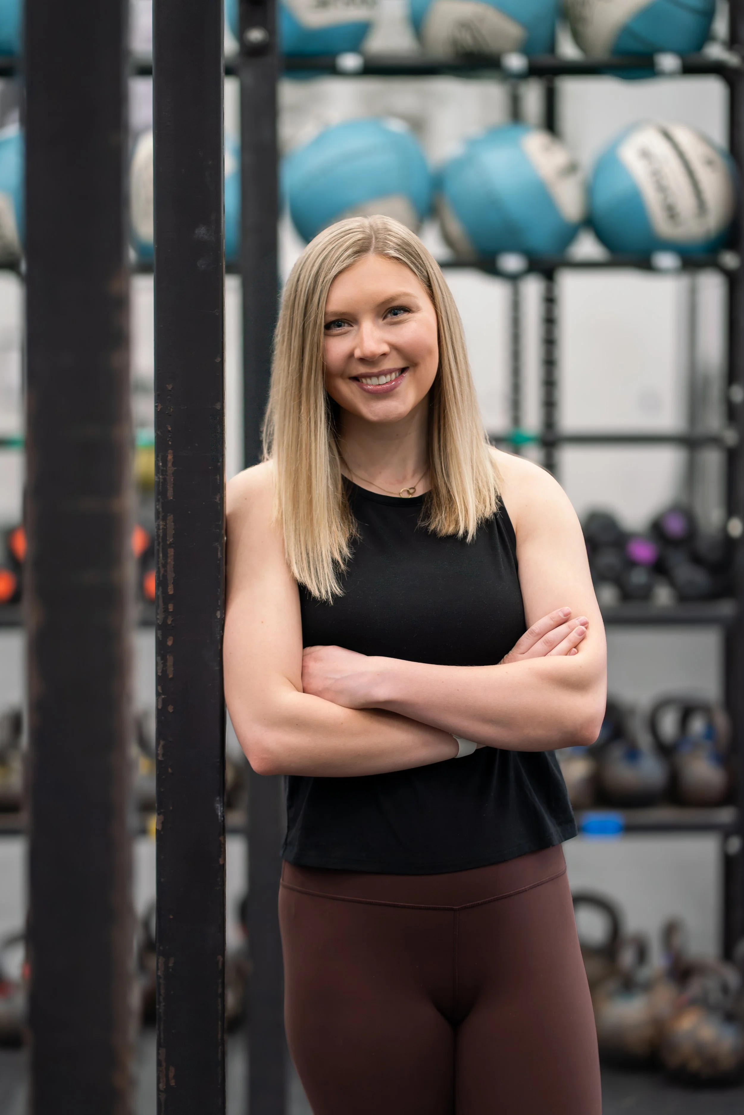 A woman with long blonde hair smiling at the camera, standing with arms crossed in a gym, with kettlebells on a rack in the background.
