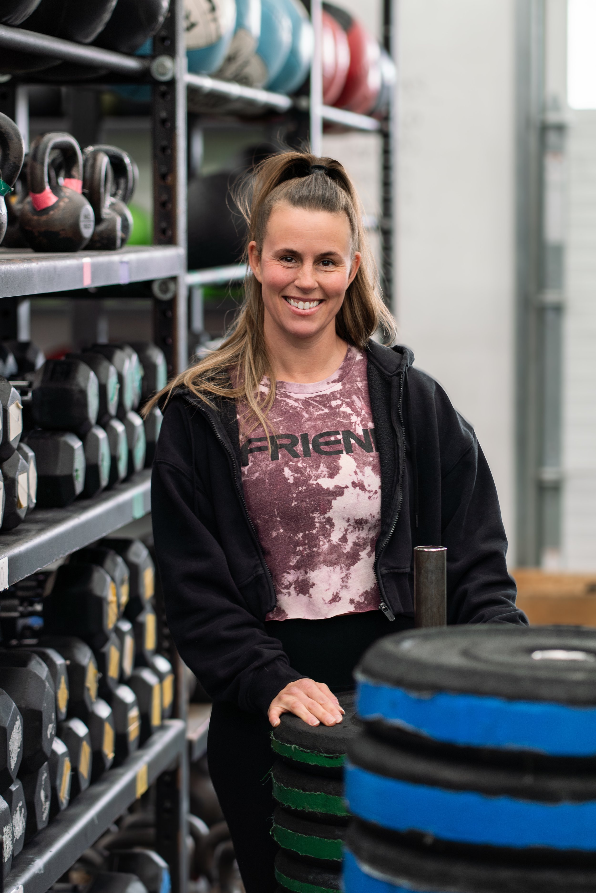 A woman standing in a gym surrounded by weightlifting equipment, smiling, with her hand on a stack of weight plates.