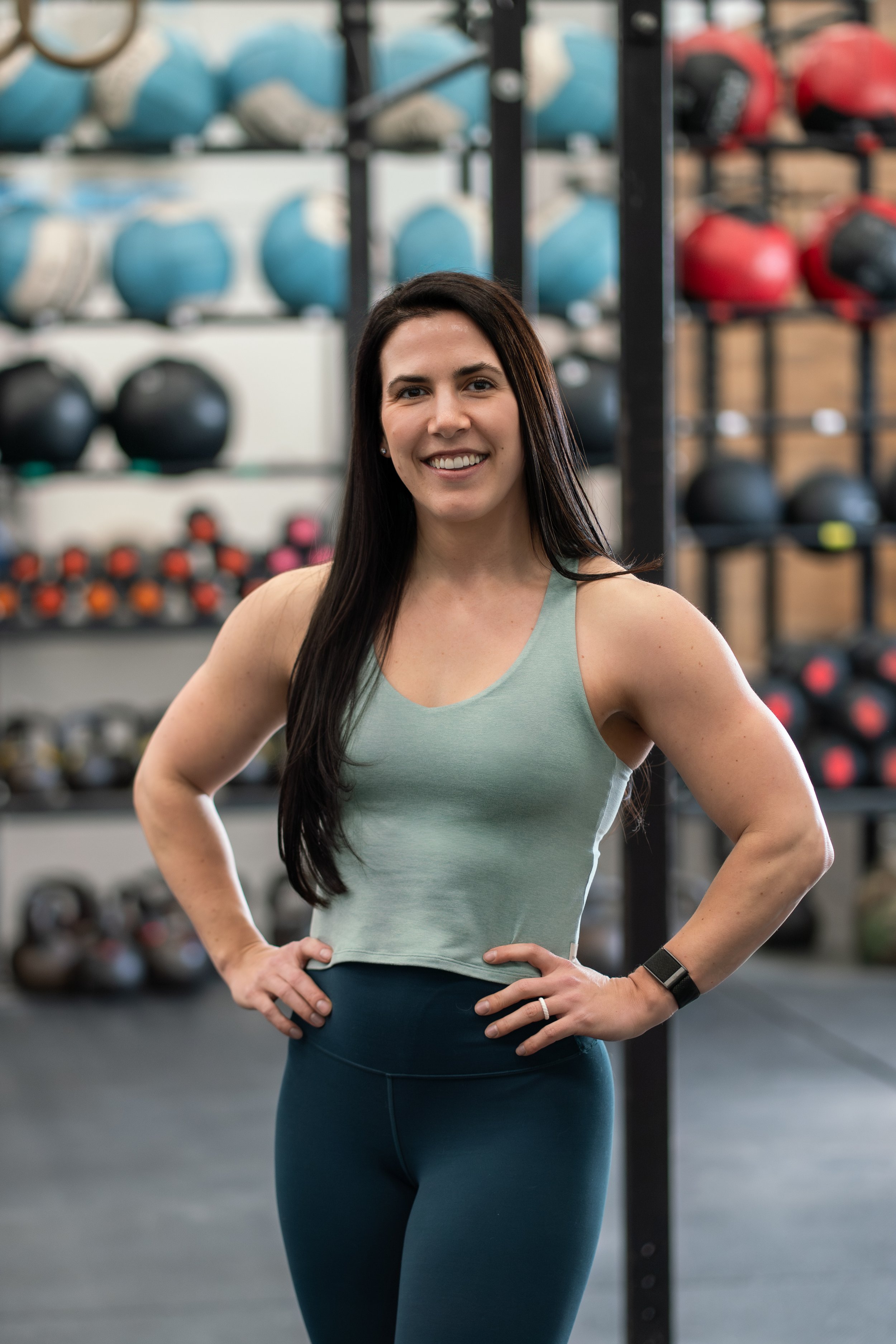 A woman with long dark hair smiling in a gym, wearing a light green tank top and dark athletic pants. She has her hands on her hips and stands in front of a wall of colored medicine balls.