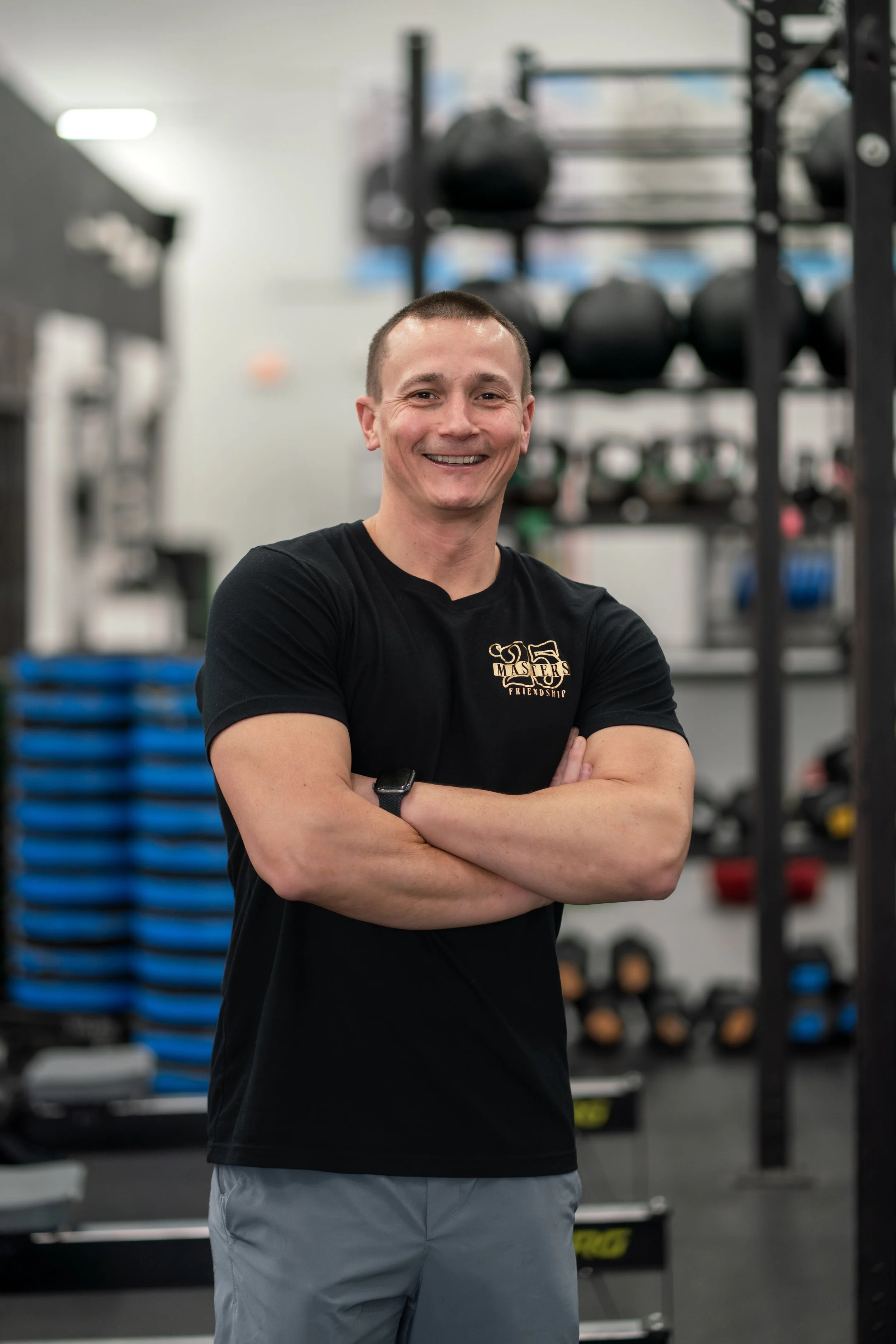 A smiling man with short hair in a gym, wearing a black T-shirt and gray shorts, standing with arms crossed, in front of weightlifting equipment and blue exercise mats.