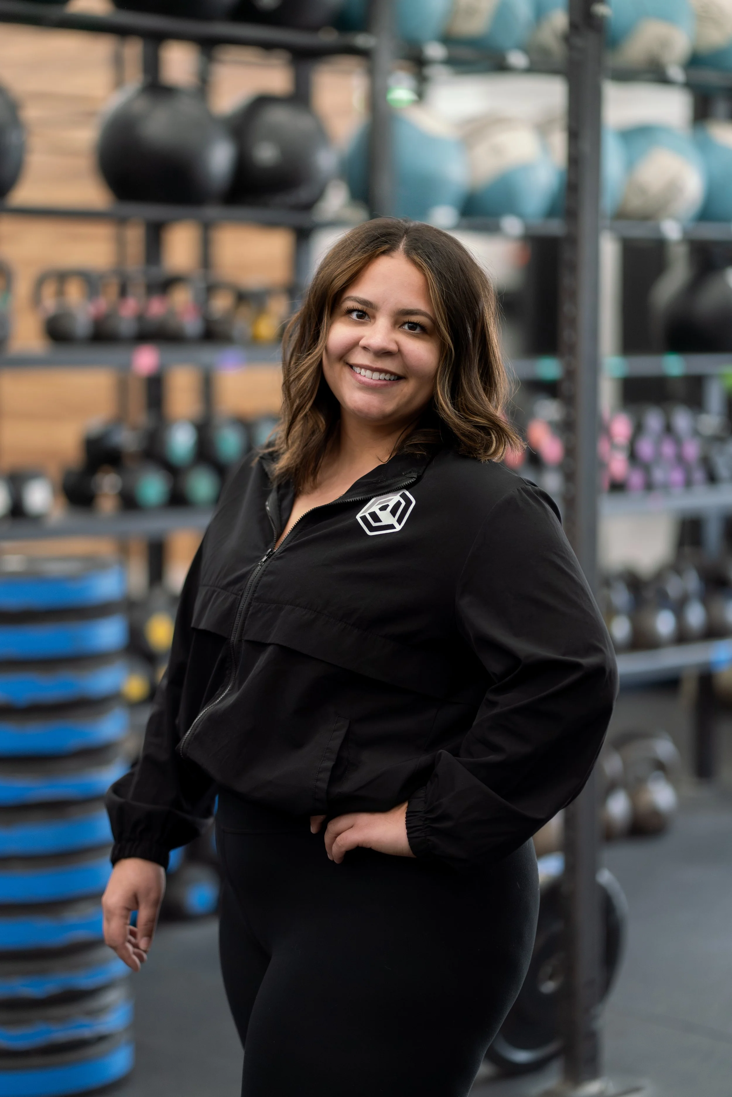 A woman smiling in a gym, standing in front of shelves with kettlebells.