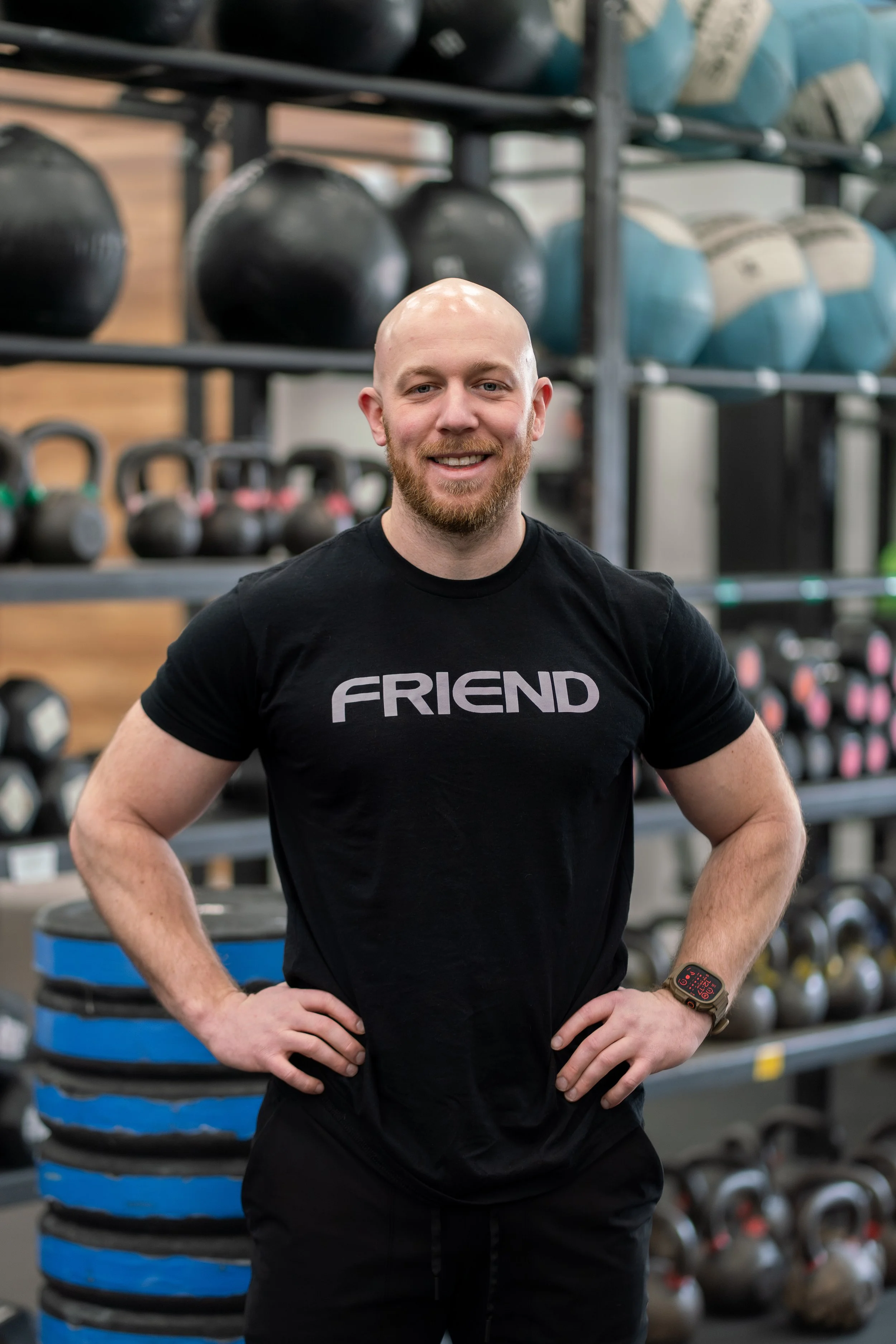 A man with a beard and a bald head smiling in a gym, wearing a black t-shirt with the word 'FRIEND' on it, standing in front of organized gym equipment including kettlebells, medicine balls, and stacked weight plates.