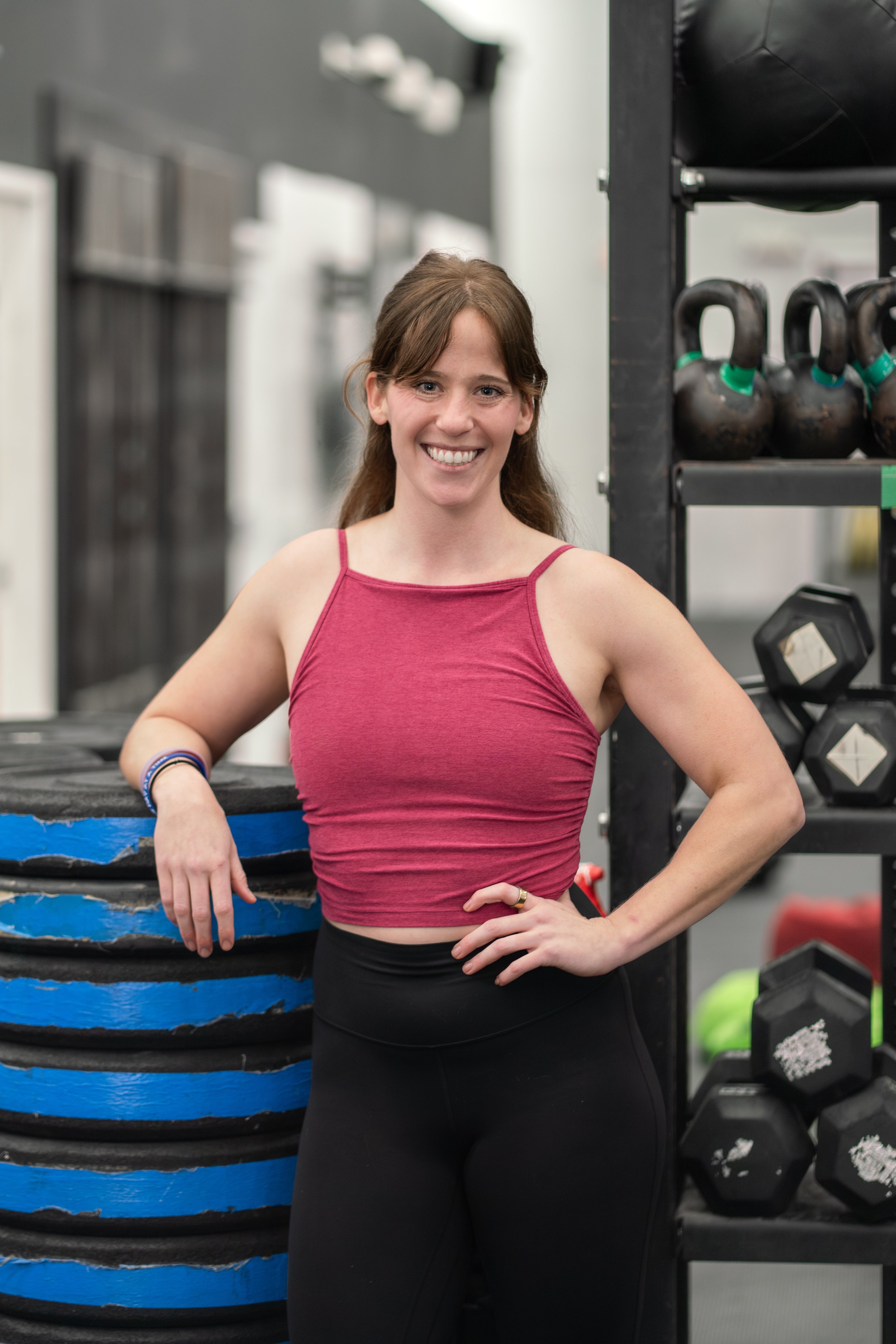 A woman with brown hair, wearing a pink sleeveless top and black leggings, smiling in a gym with black kettlebells and weight plates in the background.