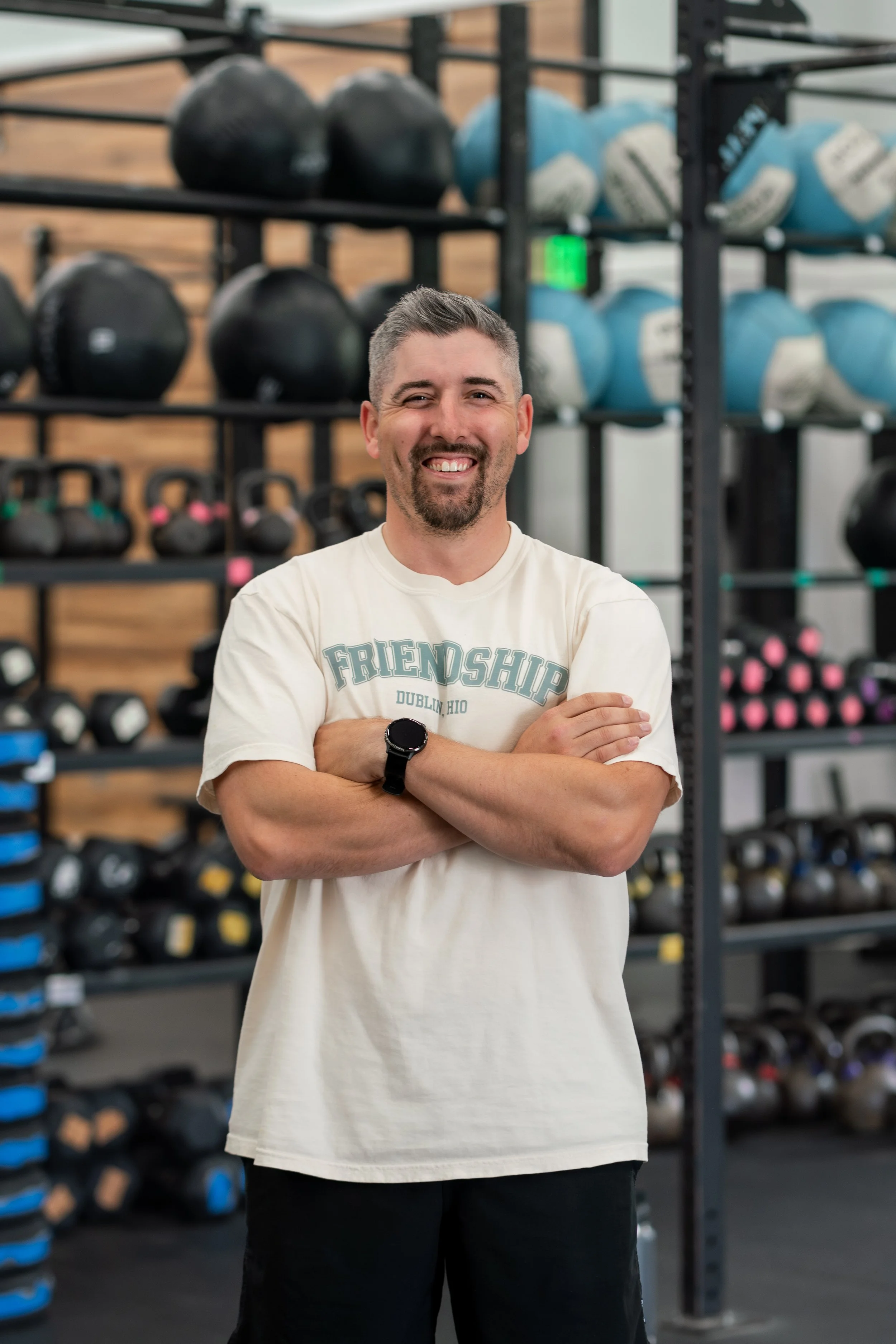 A man smiling with arms crossed, standing in a gym with black kettlebells and blue weights on shelves in the background.