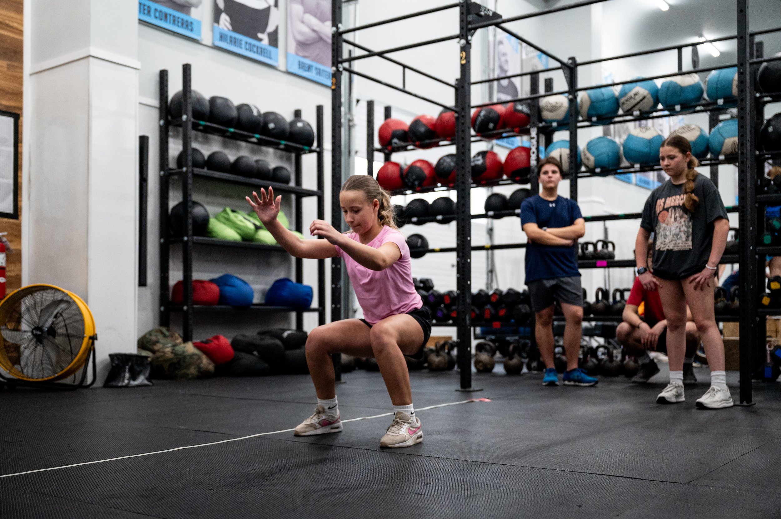 A young girl in a pink shirt and black shorts is exercising indoors on a black gym floor, jumping over a rope. Two other children are standing nearby, watching her. The gym has racks of medicine balls, a fan, and posters on the wall.