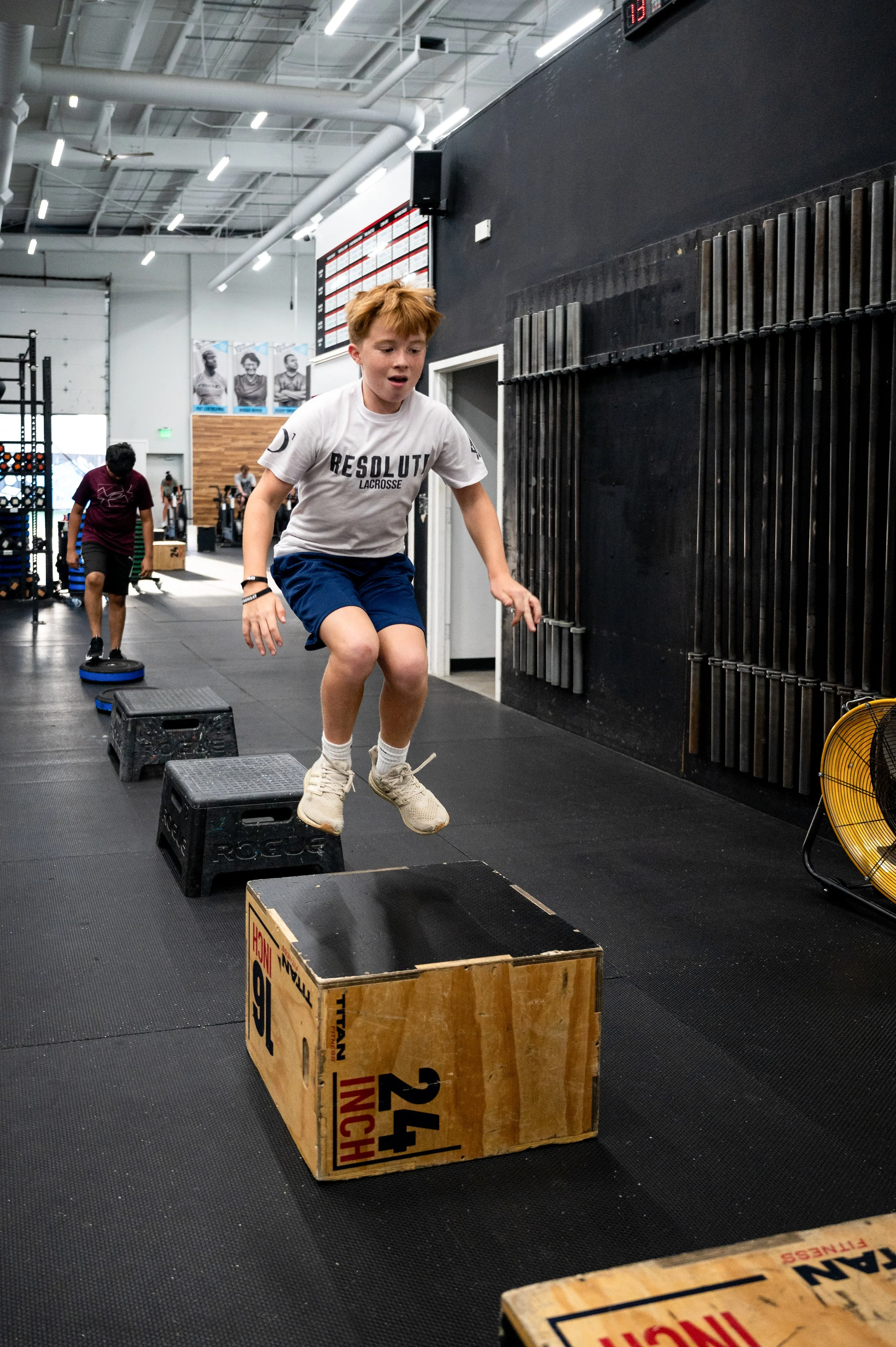 A young boy with red hair in athletic wear jumps onto a plyometric box in a gym with black flooring, while other children are exercising in the background.
