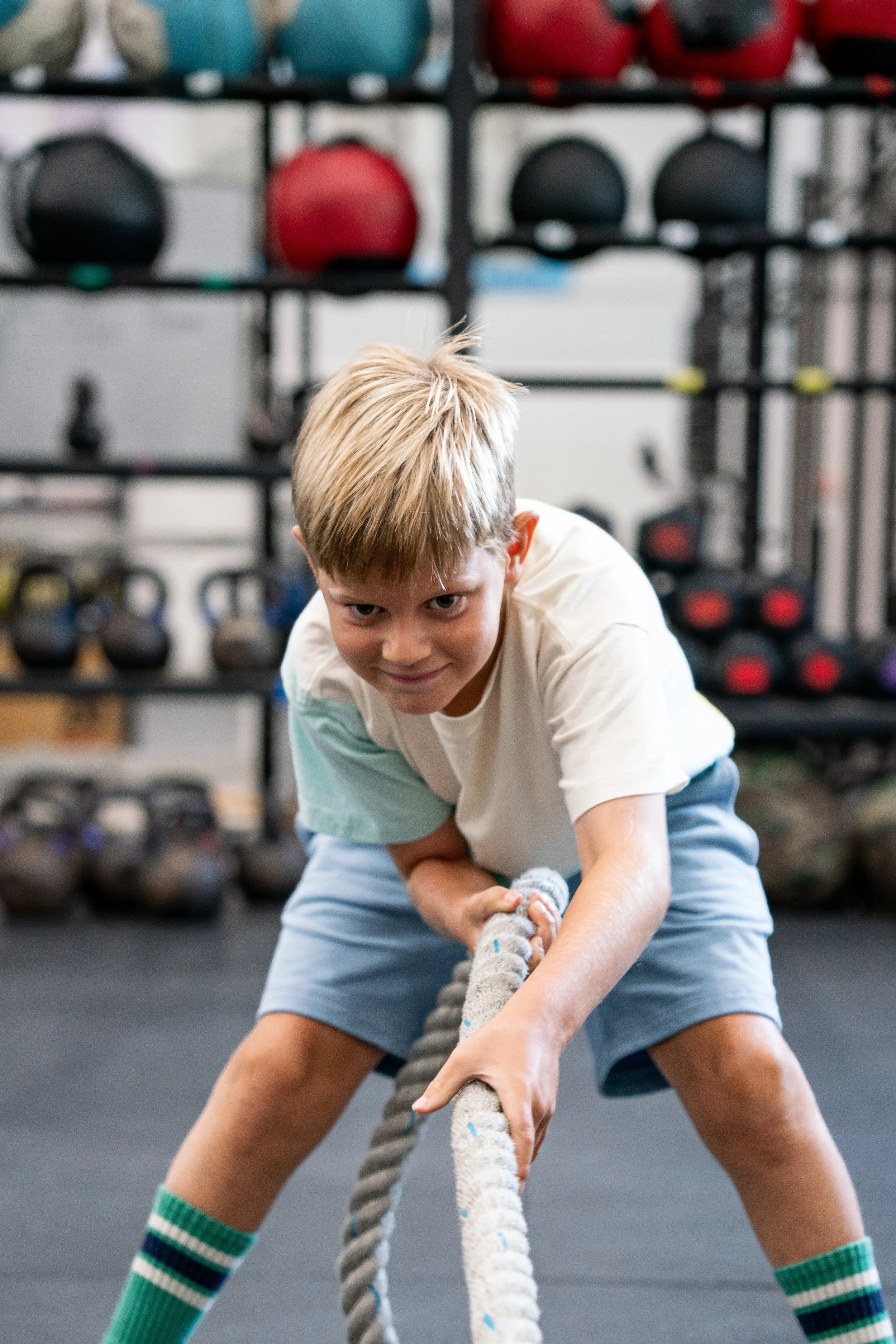 A young boy with blond hair in a gym is engaging in a tug-of-war game, holding a thick rope with a determined expression. In the background, there are shelves with various colored kettlebells.