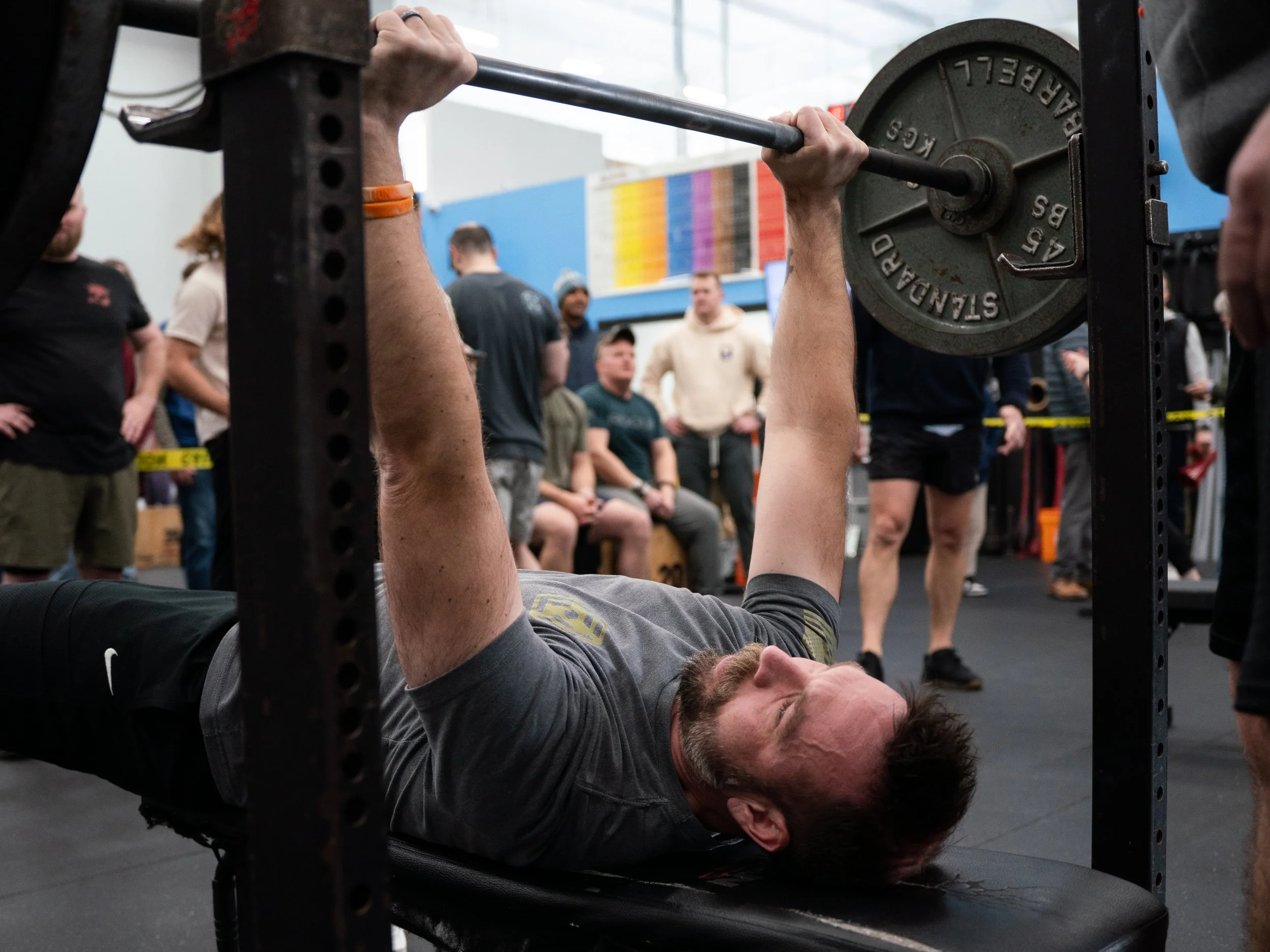 A person is lying on a weight bench, lifting a barbell with red and black weights while a trainer watches and assists. The trainer is wearing a gray T-shirt with the word 'LIFT.' printed on it. The gym has various weight plates and equipment in the background.