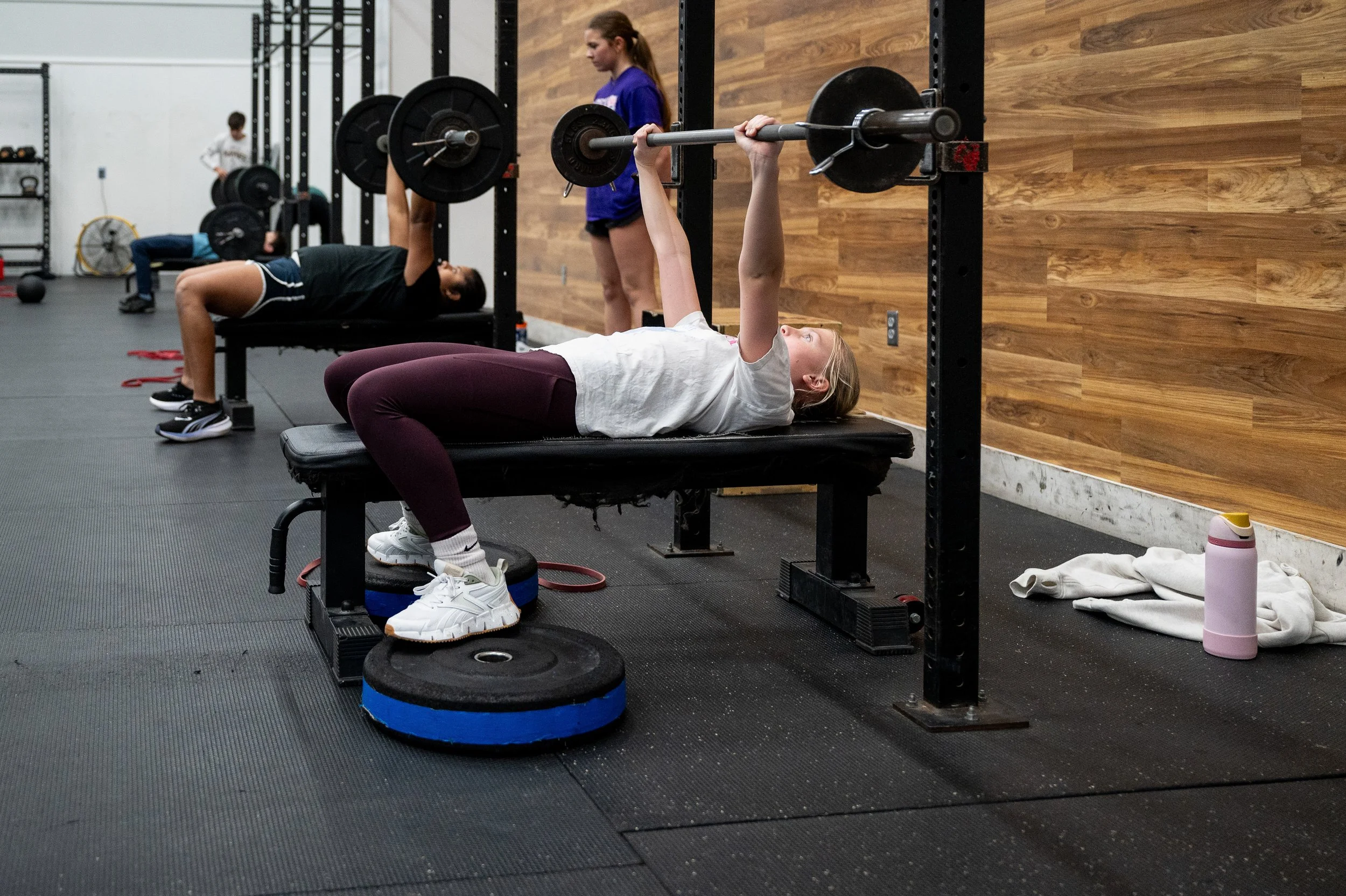 Young girl lying on a bench press, lifting a barbell in a gym.