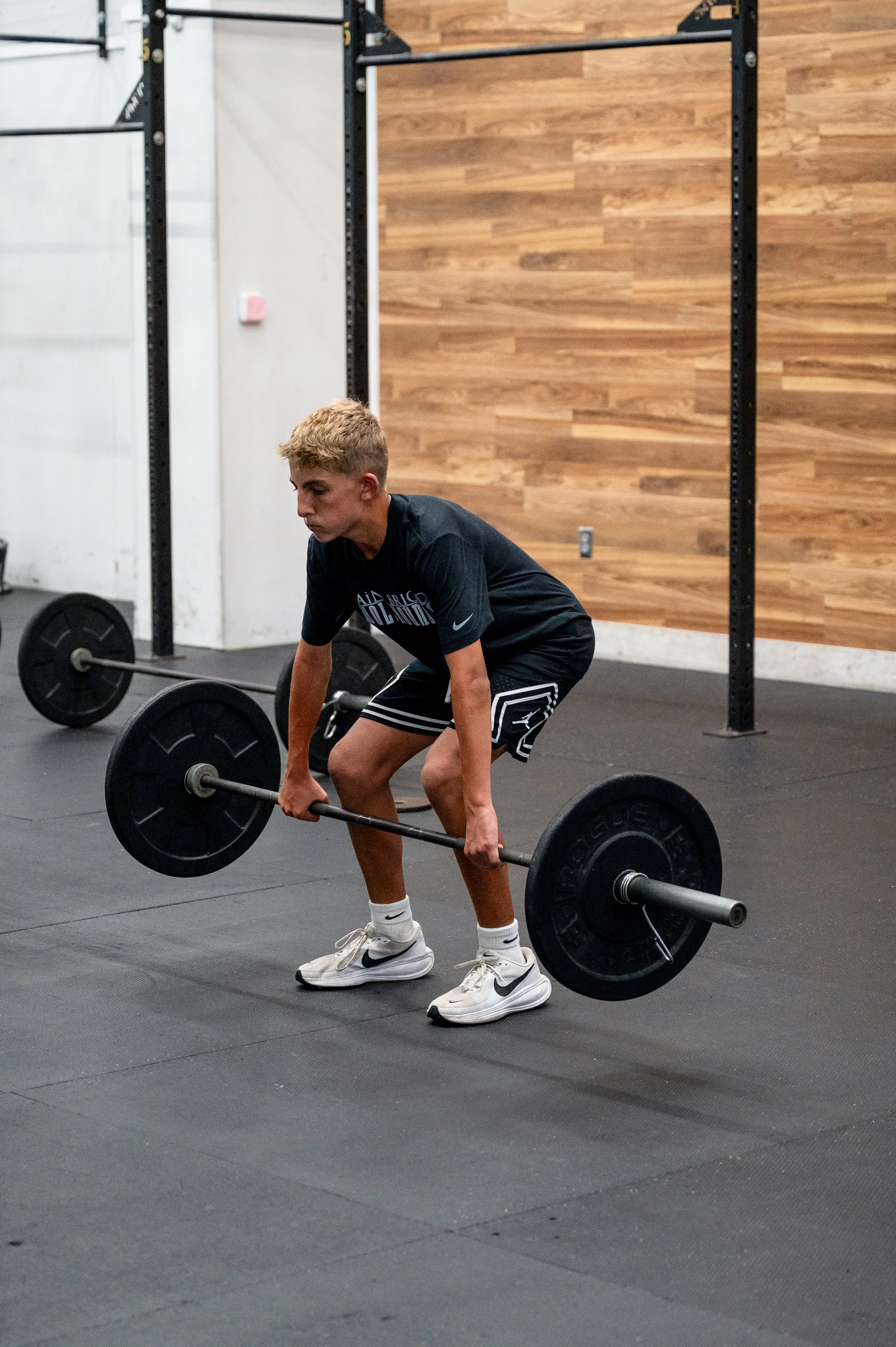 Young man in athletic clothes lifting a barbell with weights in a gym with wood paneling on the wall.