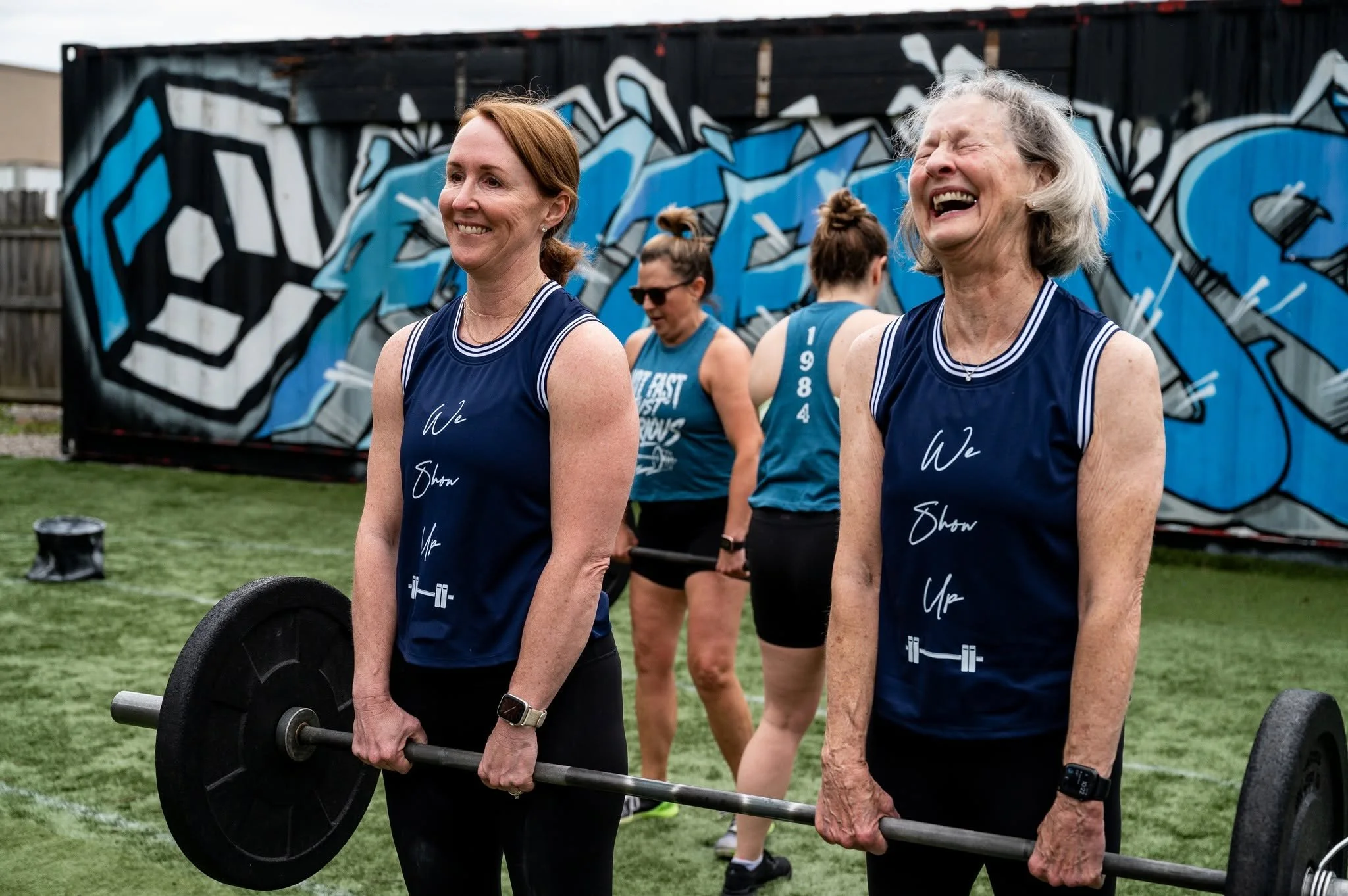 Two women of different generations smile while holding barbells during an outdoor group fitness class, with other participants exercising in the background and a colorful blue-and-black mural behind them.