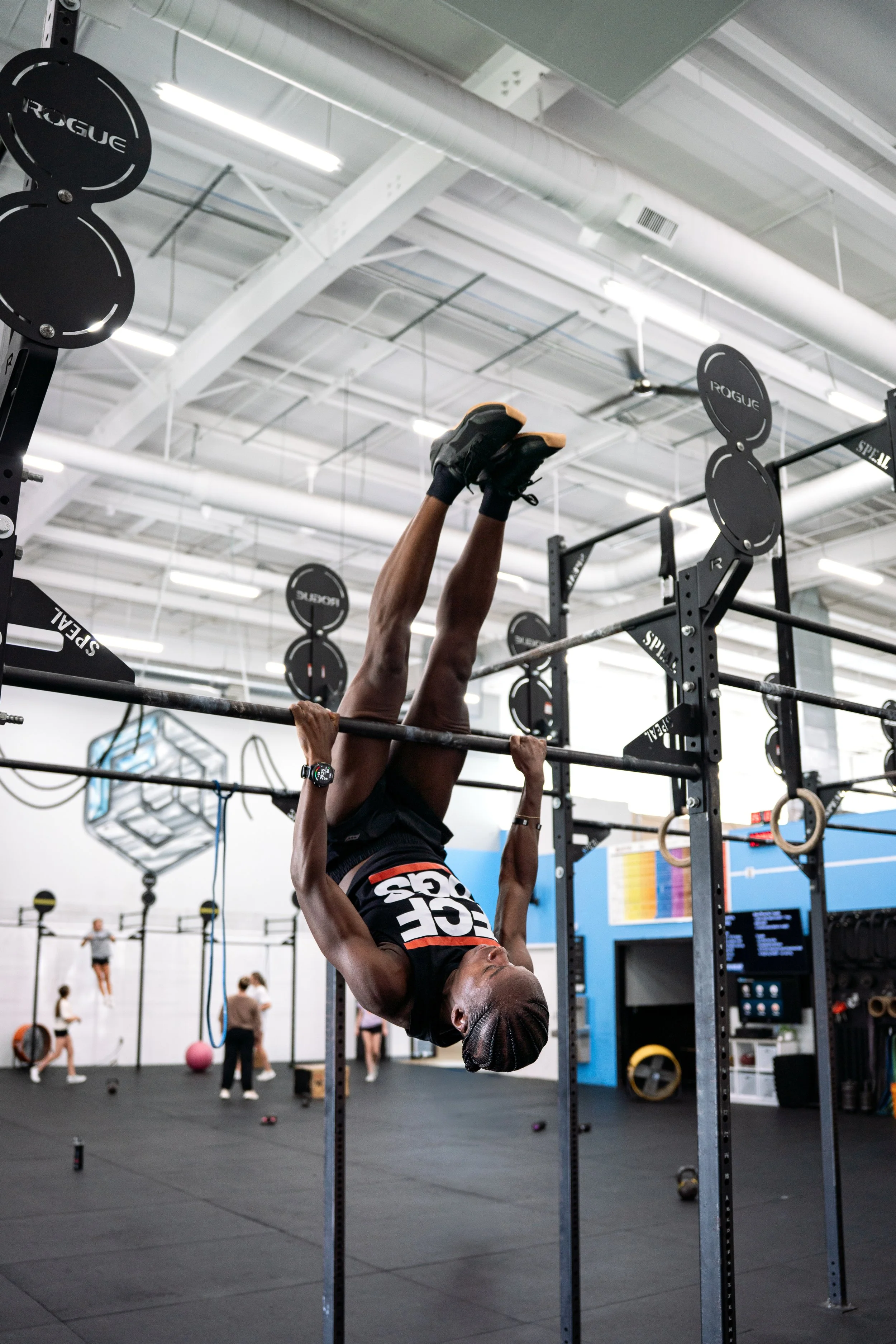 A person doing an inverted exercise on a pull-up bar at a gym surrounded by other people and workout equipment.