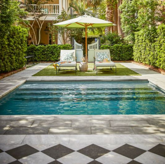 A backyard swimming pool with two lounge chairs under a large umbrella, surrounded by lush greenery and a white fence.