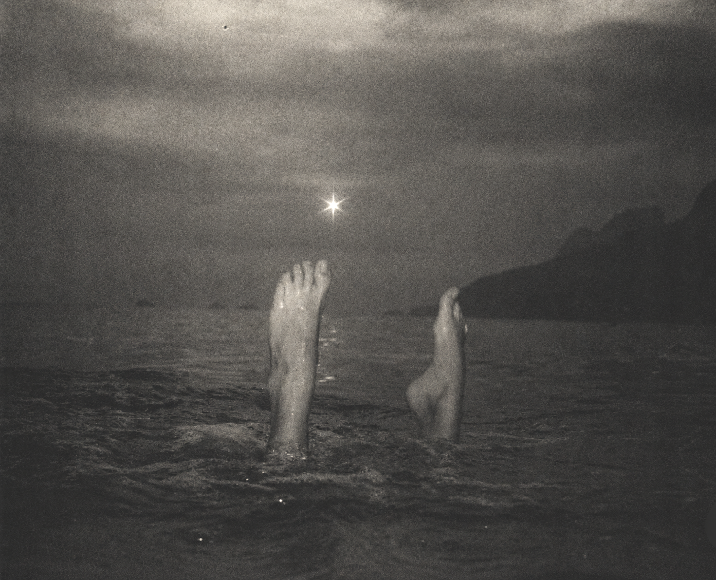 A person floating in the water with legs and feet visible above the surface, at night, with the moon shining in the sky and a distant shoreline or island silhouette.