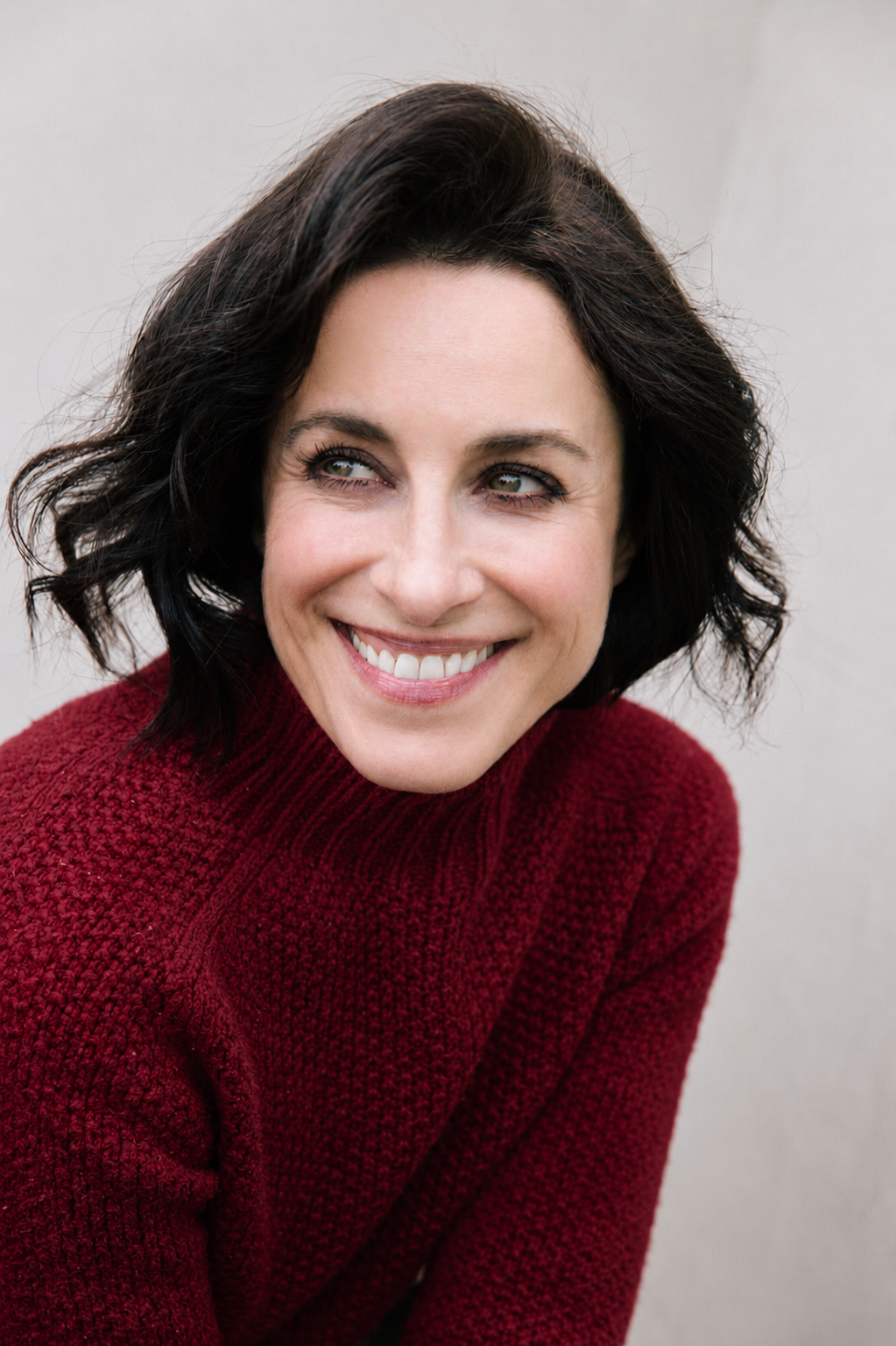 A woman with short dark hair wearing a red sweater, smiling and looking to her left, against a light gray background.