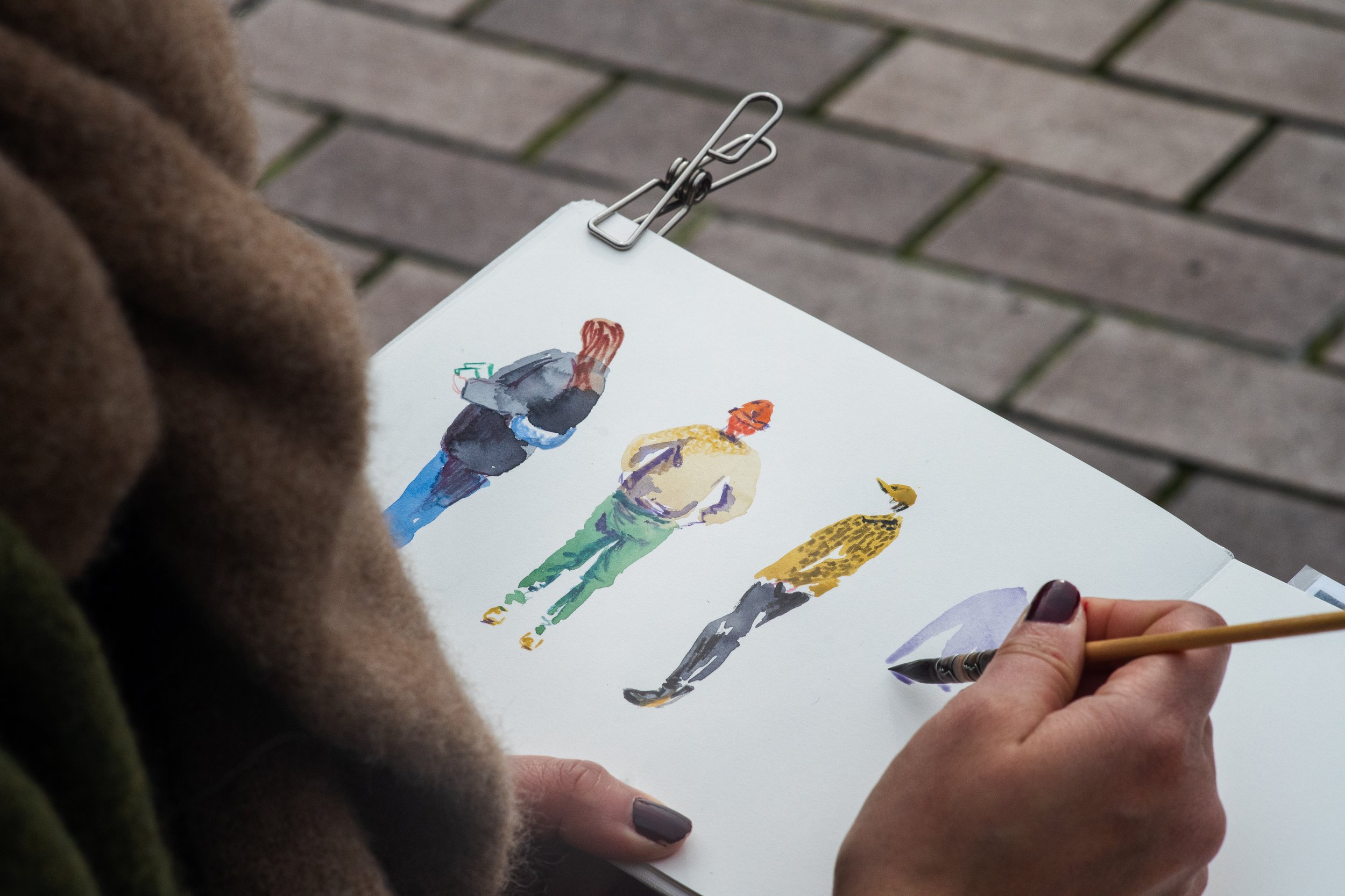 Une personne dessine ou peint des silhouettes de personnes en utilisant de l'aquarelle sur un carnet. La scène se déroule sur un sol pavé.