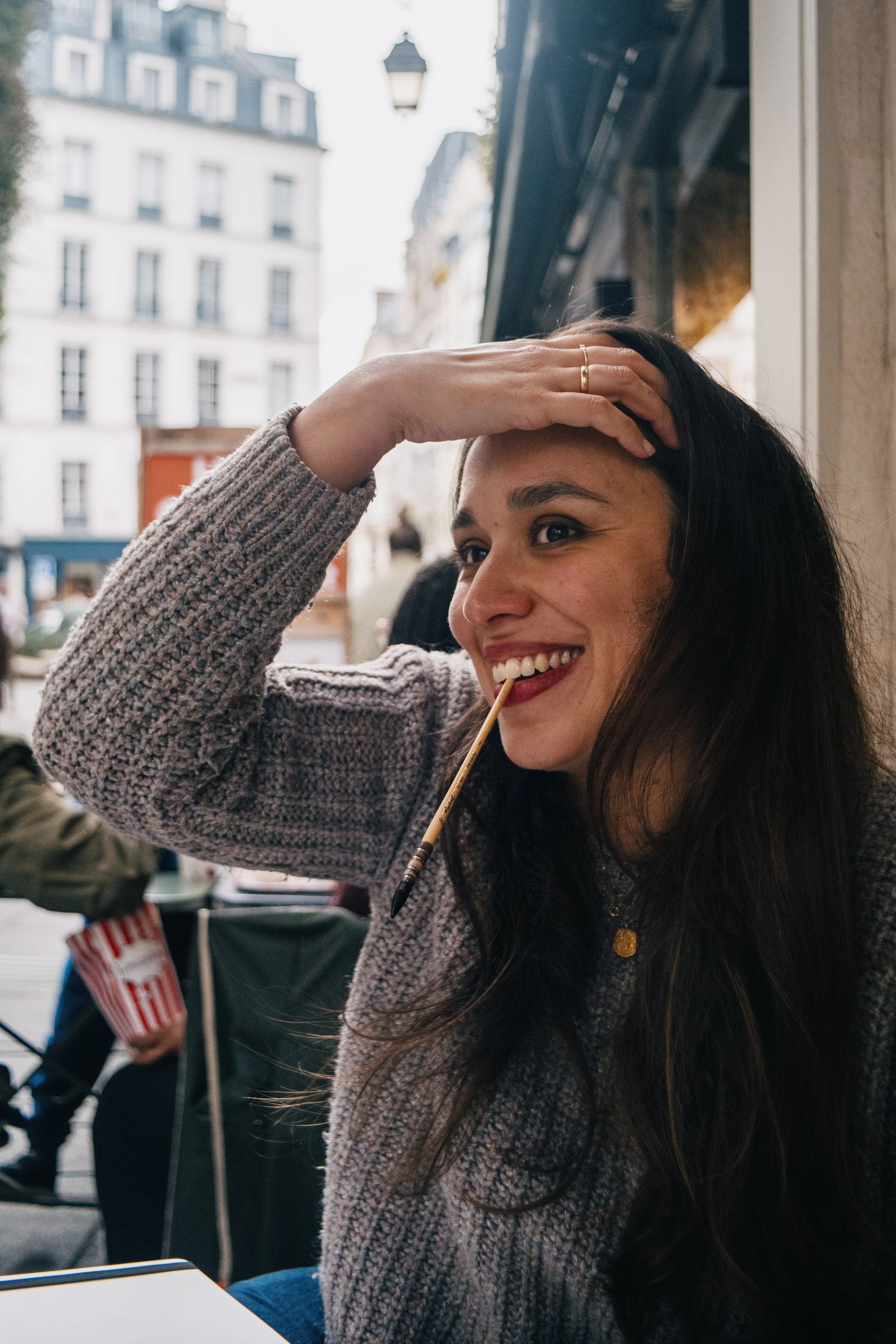 Une jeune femme souriante avec des cheveux longs, portant un pull beige, siège dans un café en tenant un cure-dent avec une glace. Elle a la main sur le front, regarde à gauche, avec un décor urbain en arrière-plan.