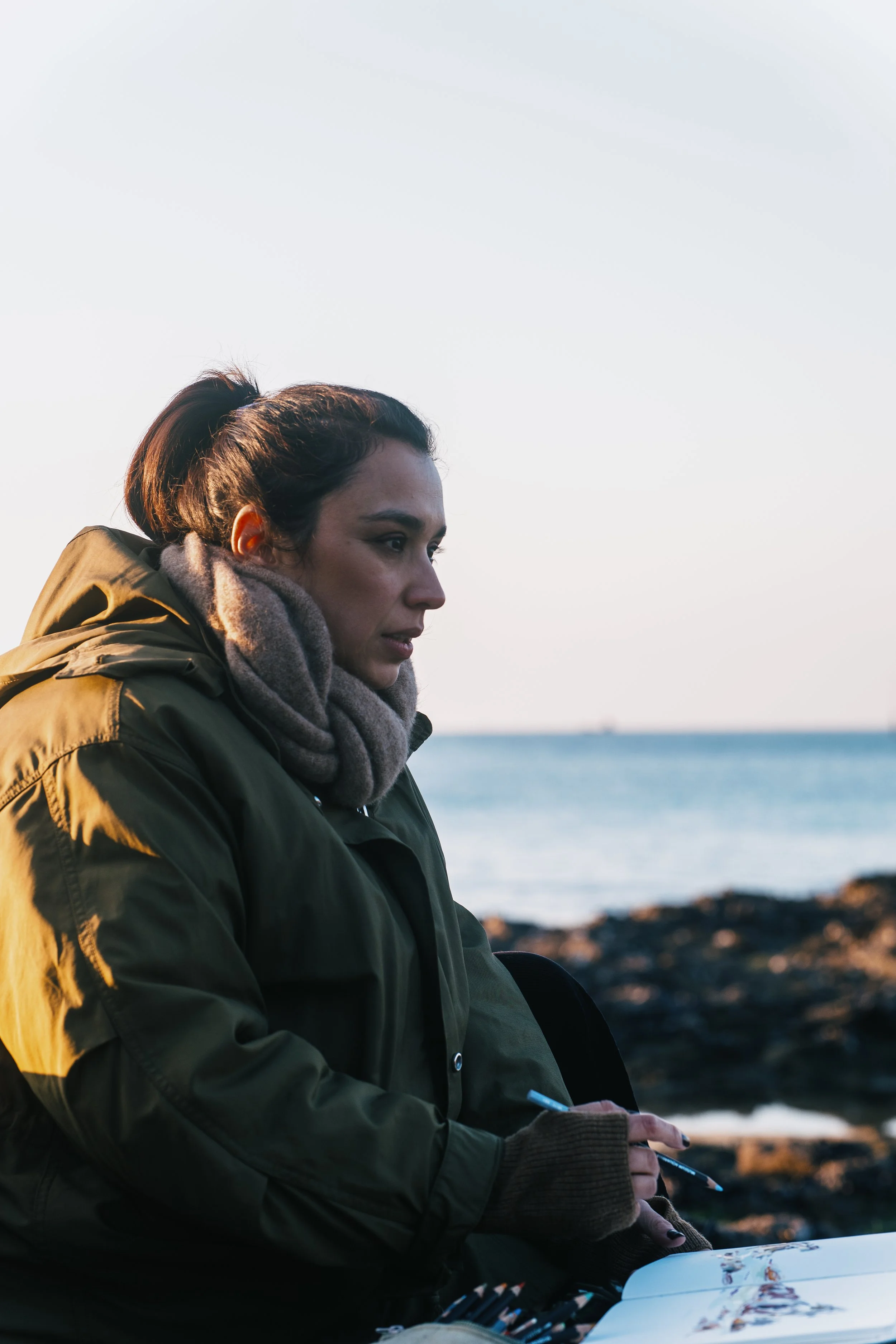 Femme assise en plein air près de la mer, portant un manteau vert olive et une écharpe grise, en train de dessiner dans un cahier.