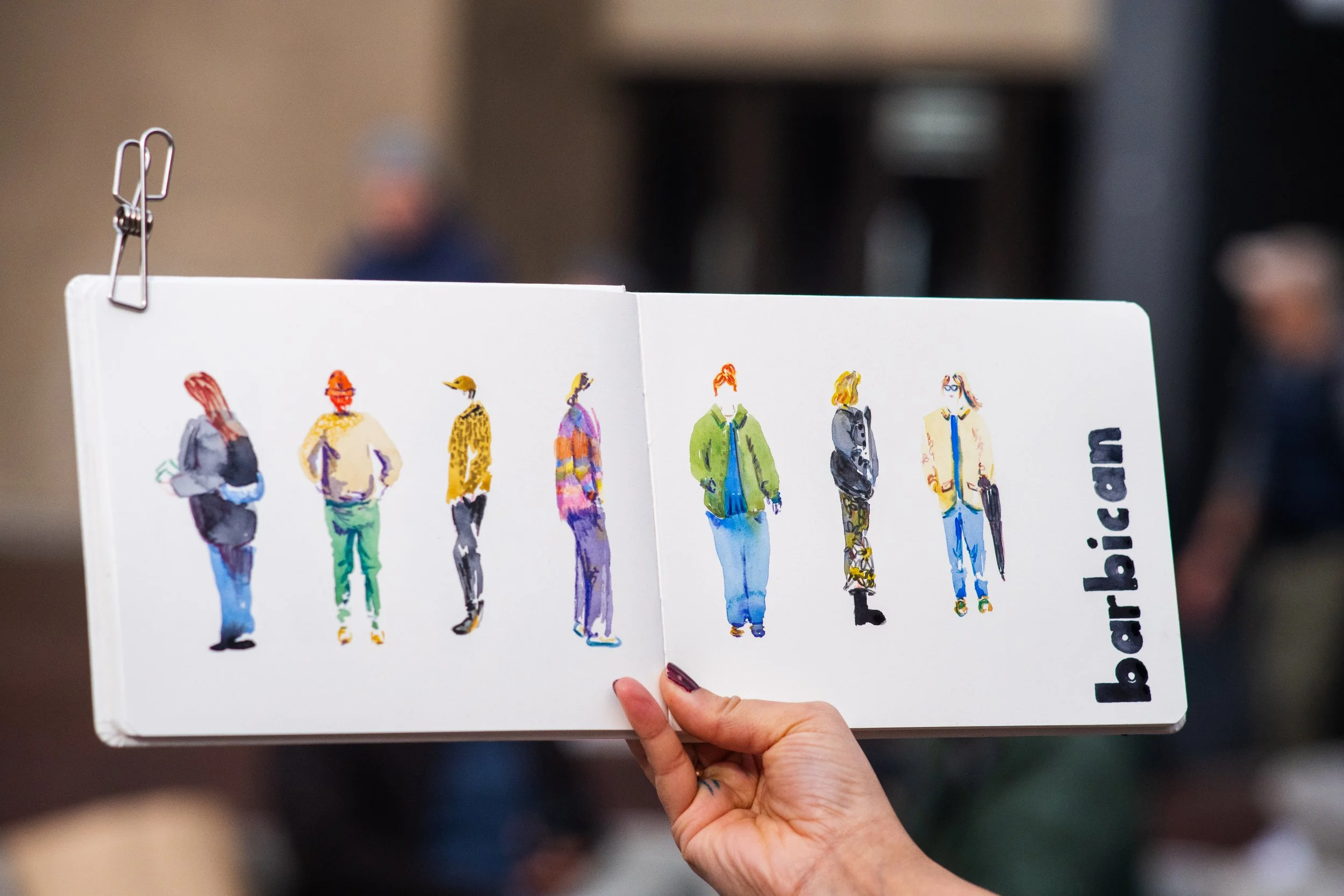 Une main tient un livre avec des illustrations de plusieurs personnes en silhouette, la page est blanche avec un texte partiel en bas à droite.