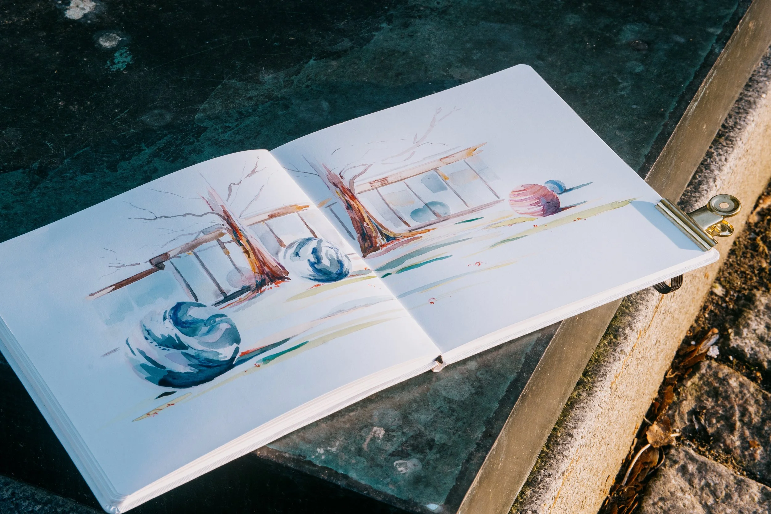 Carnet de croquis ouvert avec une peinture de nature morte, représentant des branches, des pommes et des fruits, posé sur une table en plein air.