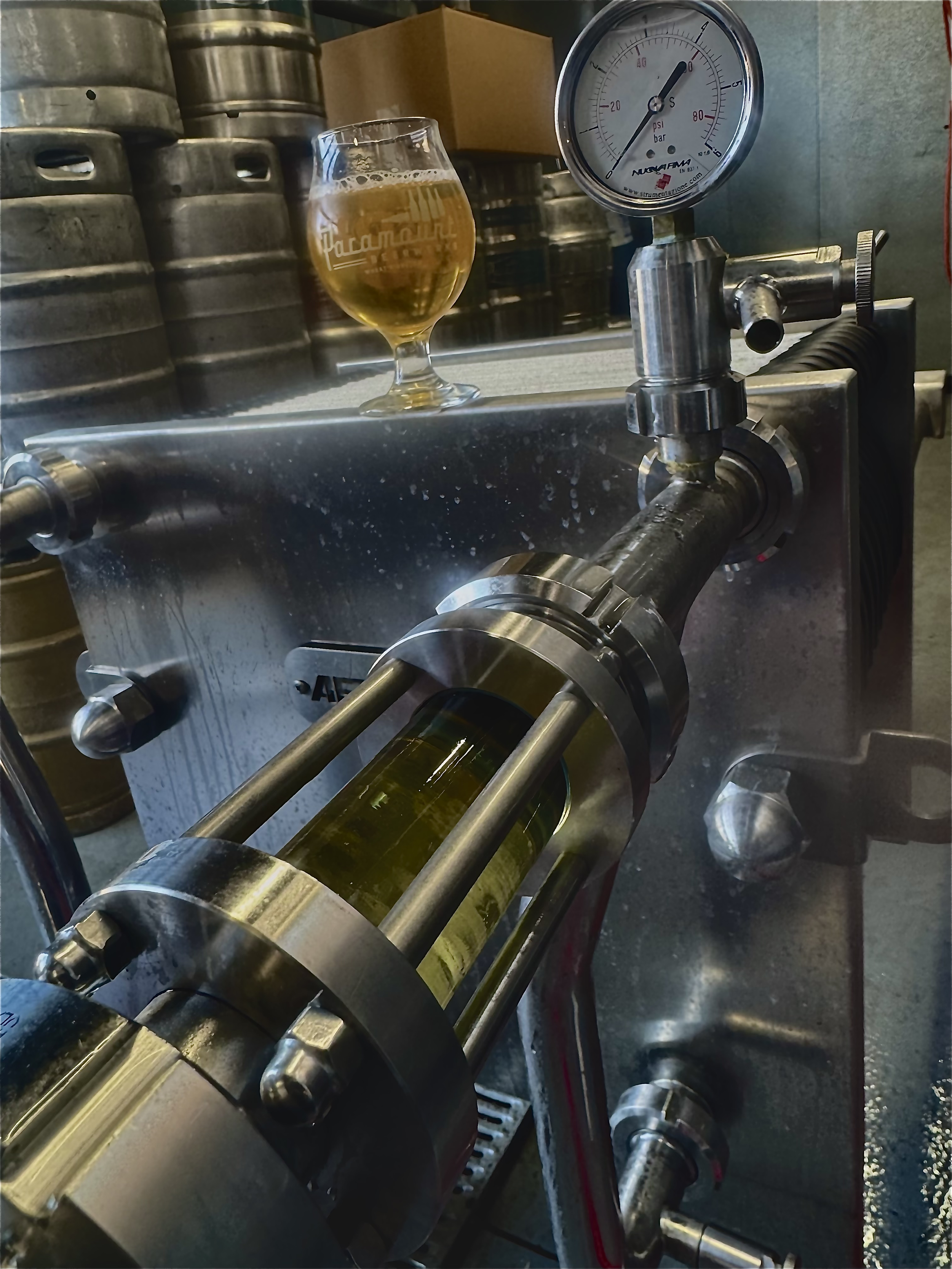 Close-up view of a metal beer can labeling machine with a cylindrical part in the foreground, a glass filled with beer on a scale, and a pressure gauge in the background.