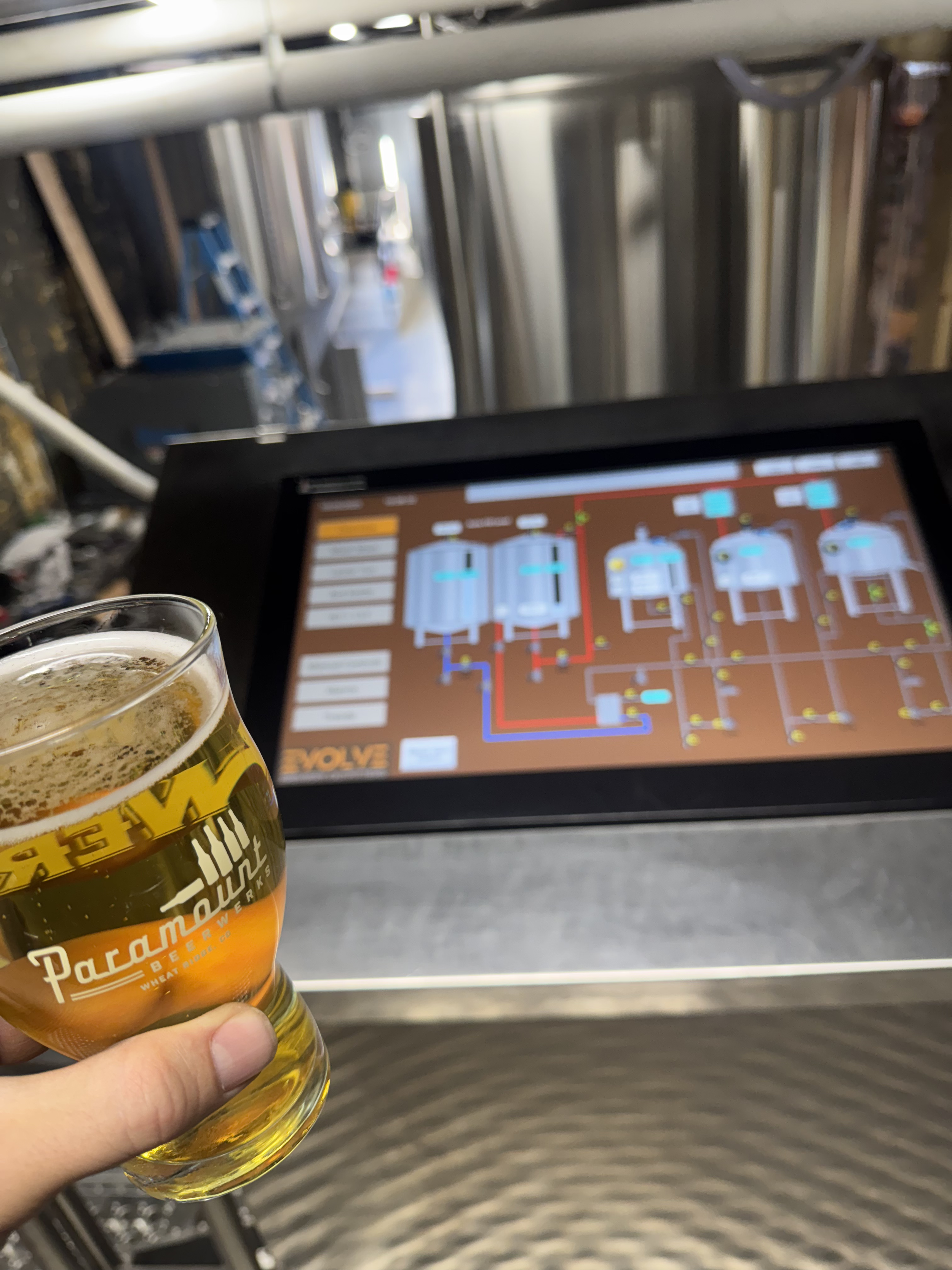 A person holding a glass of beer in front of an industrial control panel with a schematic diagram.