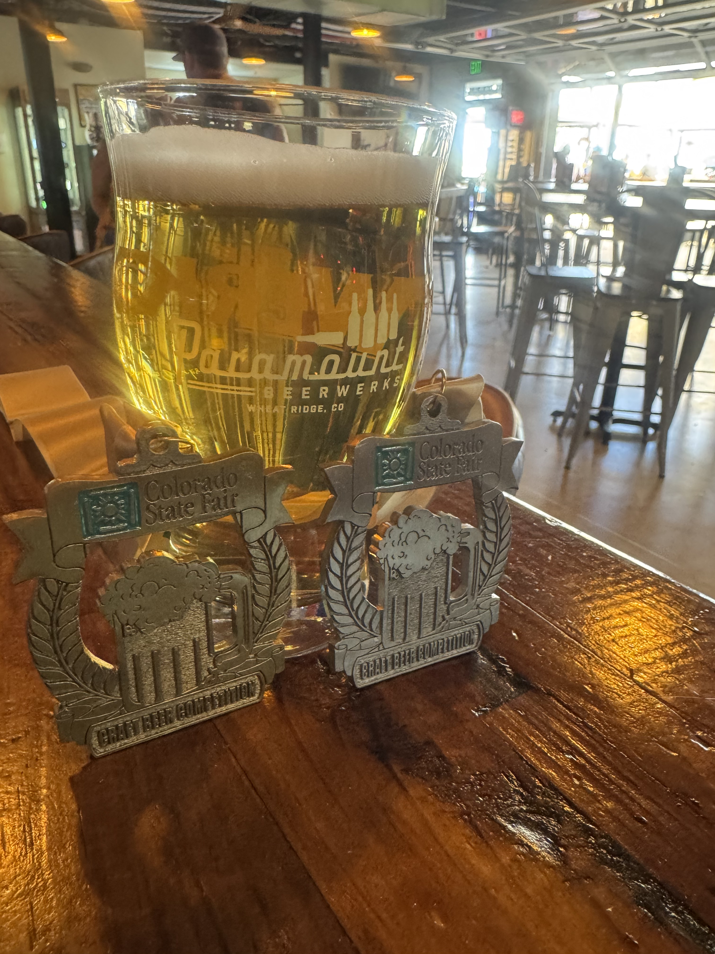 A glass of beer featuring the Paramount BeerWerks logo sits on a wooden bar next to two Colorado State Fair craft beer competition medals. The background shows the interior of the establishment, including tables and chairs.