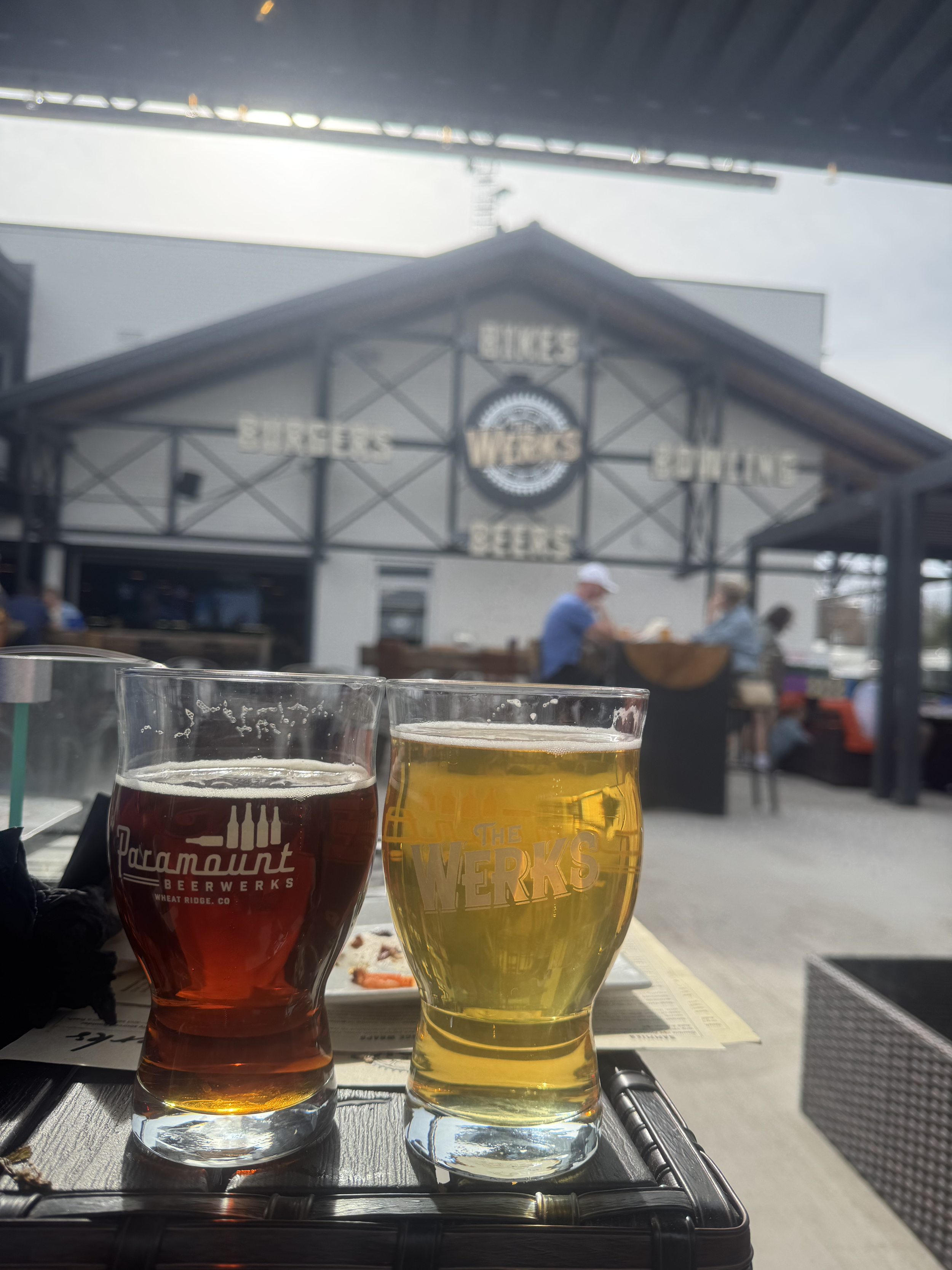 Two glasses of beer, one amber and one yellow, on a tray at an outdoor restaurant. In the background, there are people dining, a building with a sign that says 'THE WERKS,' and a bright sky.