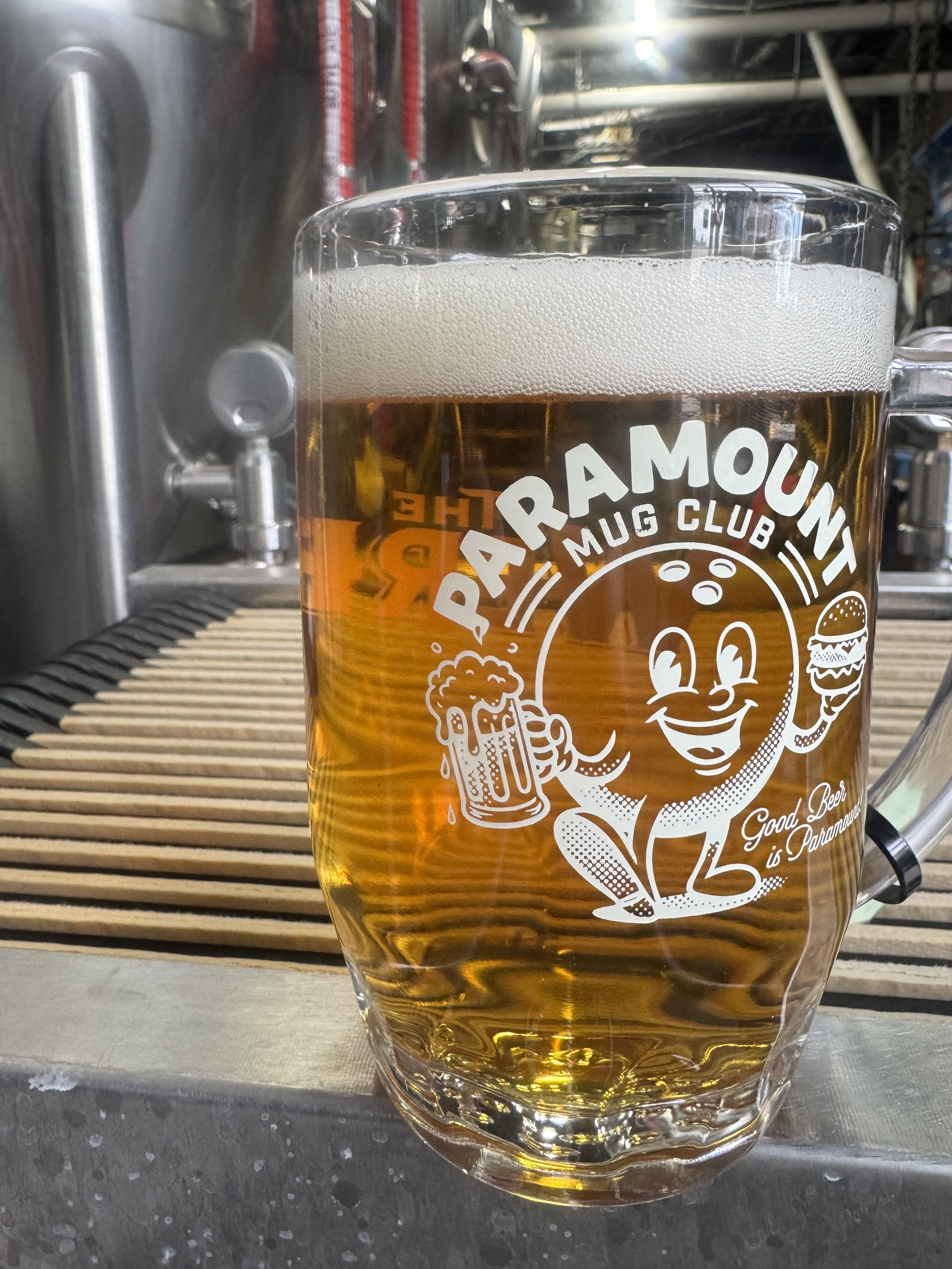 A full mug of beer sits on a stainless steel bar, with brewing equipment visible in the background. The glass features a "Paramount Mug Club" logo depicting a cartoon bowling ball character holding a beer mug and a hamburger.