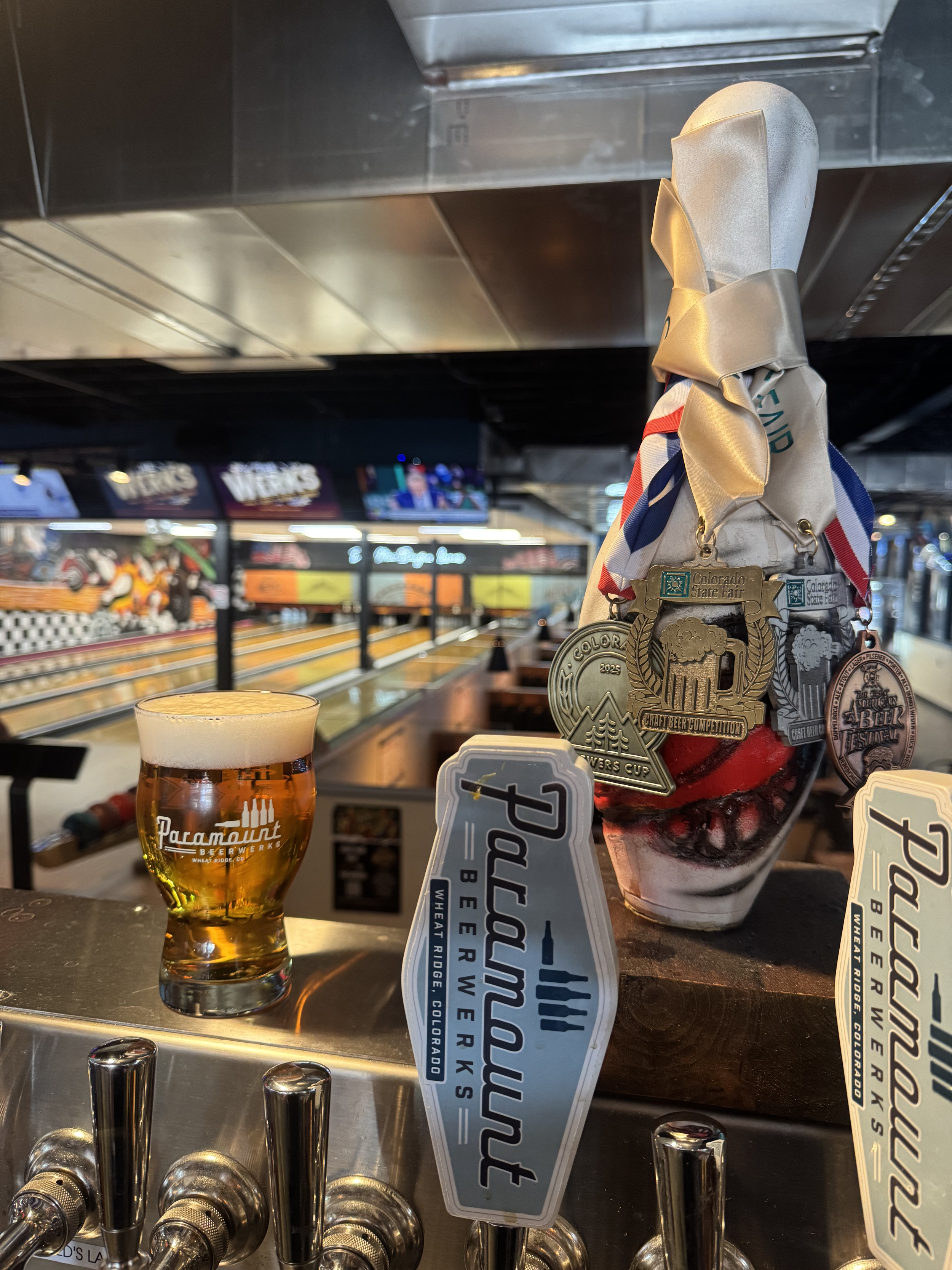A glass of beer featuring the Paramount BeerWerks logo sits on a bar next to tap handles branded with the same name. In the background, an indoor bowling alley is visible. A bowling pin decorated with ribbons and award medals stands on the counter.