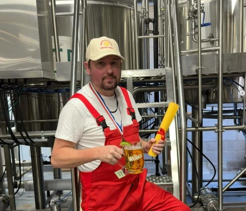 Head Brewer Chris Kirk wearing Weyermann red overalls and a branded cap, holding a beer and a certificate in the Paramount BeerWerks brewhouse.