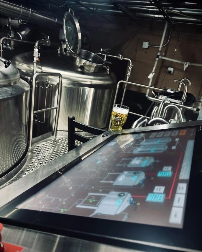 View of brewery equipment with stainless steel tanks, pipes, and a tablet displaying a brewing control interface, with a glass of beer on the counter.