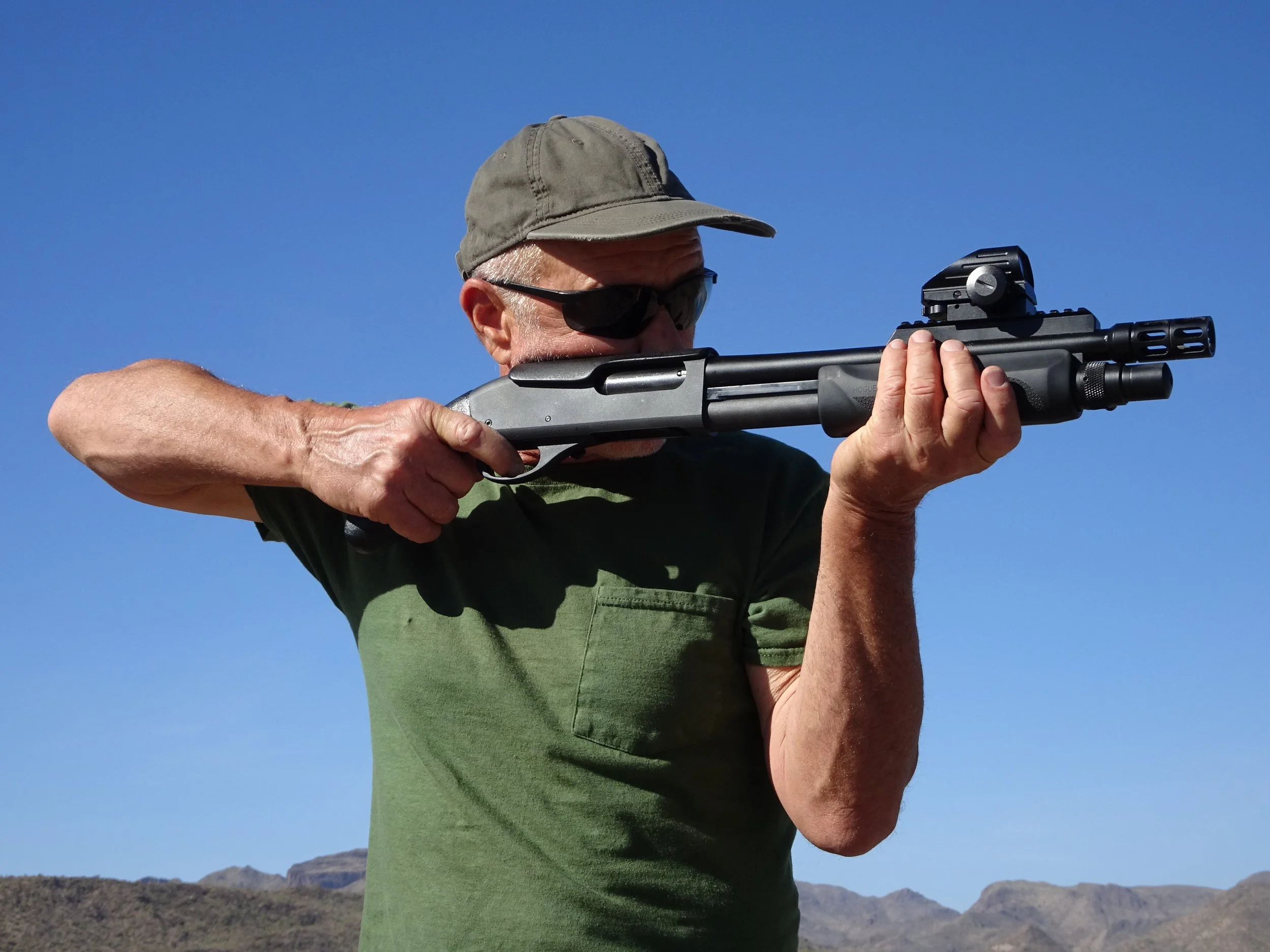 A man with a gray cap and dark sunglasses aiming a rifle, with desert landscape and mountains in the background under clear blue sky.