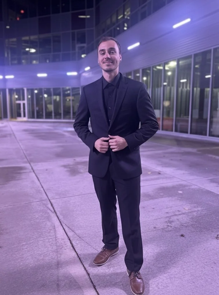 A man in a dark suit and black shirt standing outside at night in front of a modern glass building, smiling and adjusting his suit jacket.