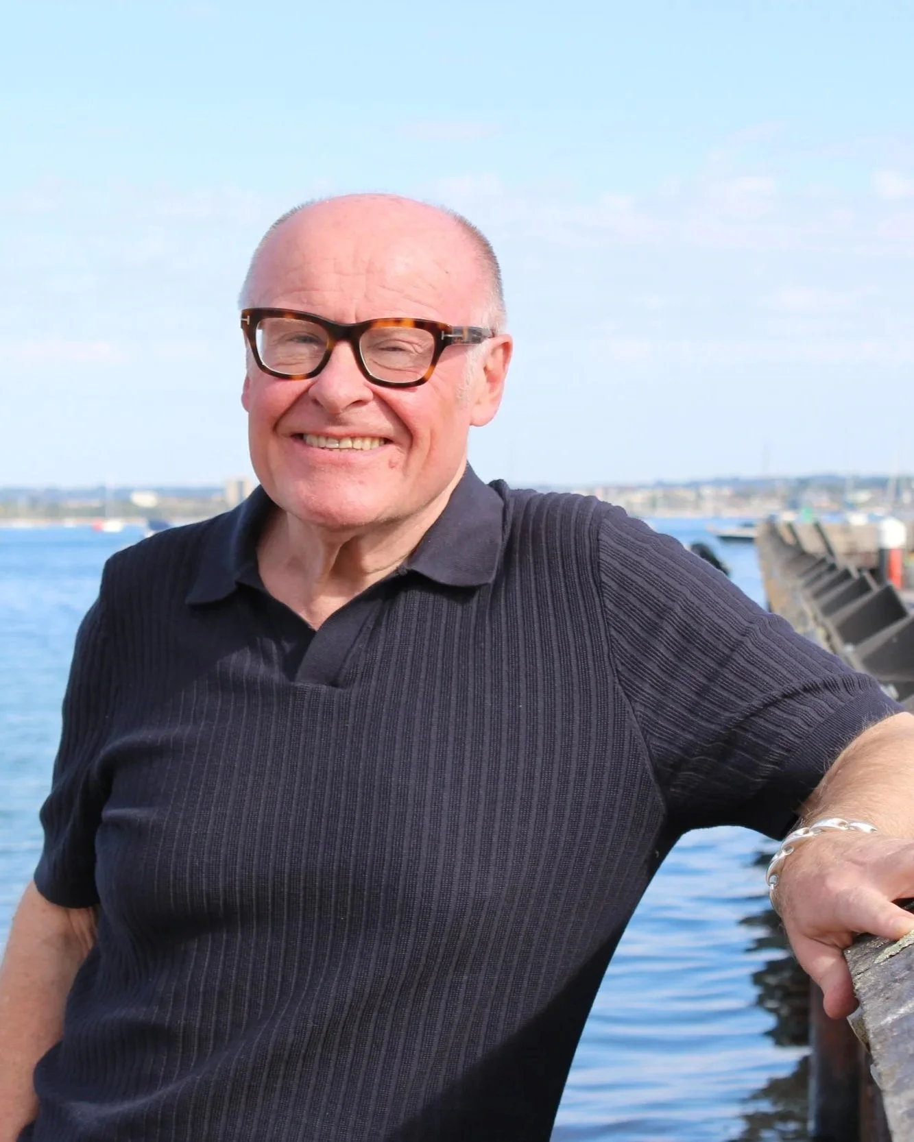 A smiling middle-aged man with glasses, wearing a black polo shirt, stands by a body of water on a sunny day with a clear sky, boats, and a distant shoreline in the background.