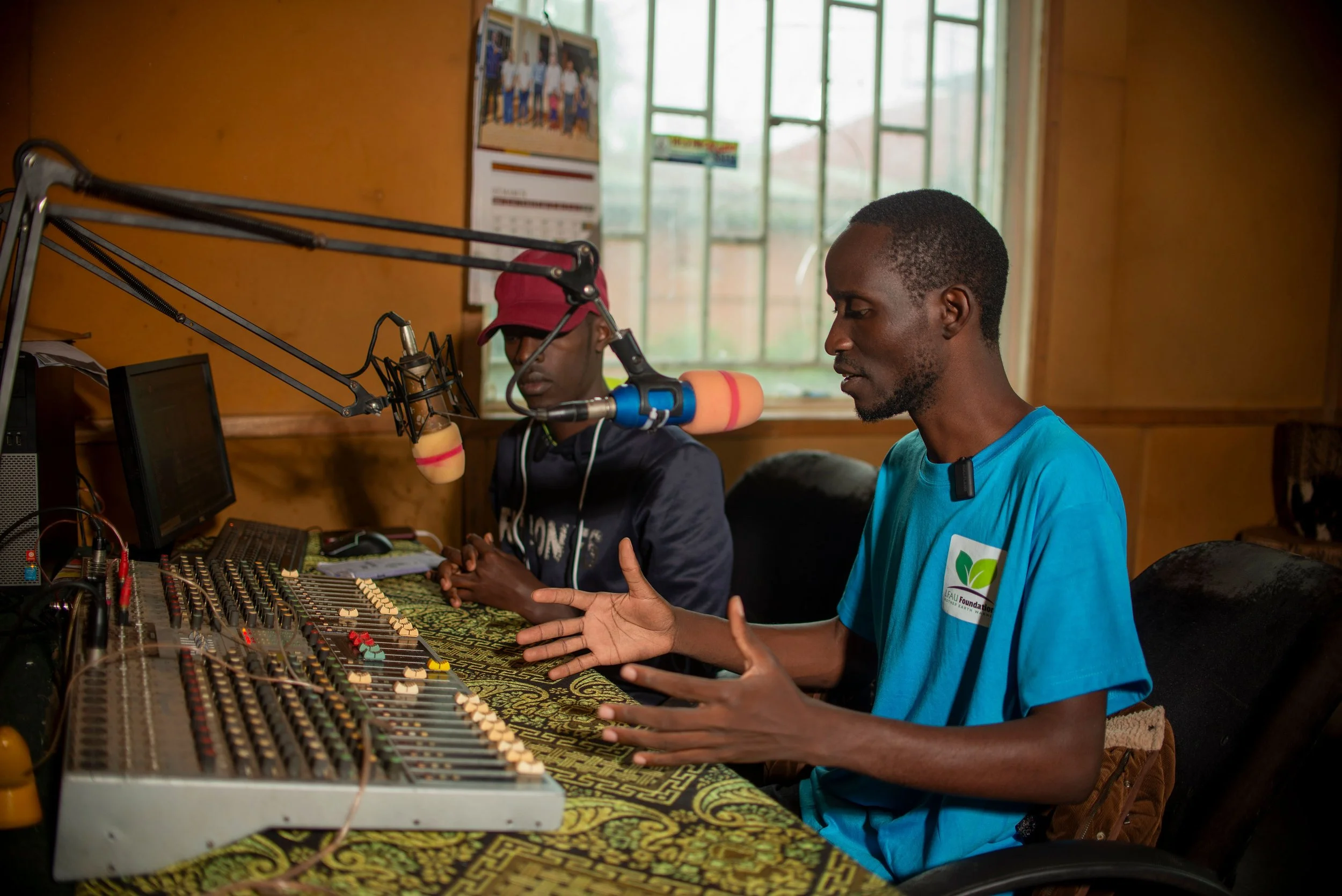 Two men are seated in front of a sound mixing console, engaging in a discussion. One man, wearing a blue shirt, is speaking passionately with hand gestures. The other man, wearing a black shirt with white text and a red cap, is looking down at a device in front of him. A microphone on a boom arm is positioned between them, and the room has wooden walls with a large window in the background letting in natural light.