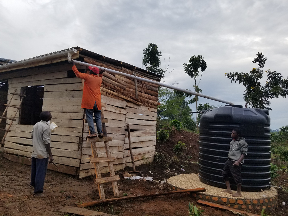 Three men near a small wooden house with a water tank being attached to the roof. One man is on a ladder, another is standing beside the house, and the third is near the water tank. The background has trees and a cloudy sky.