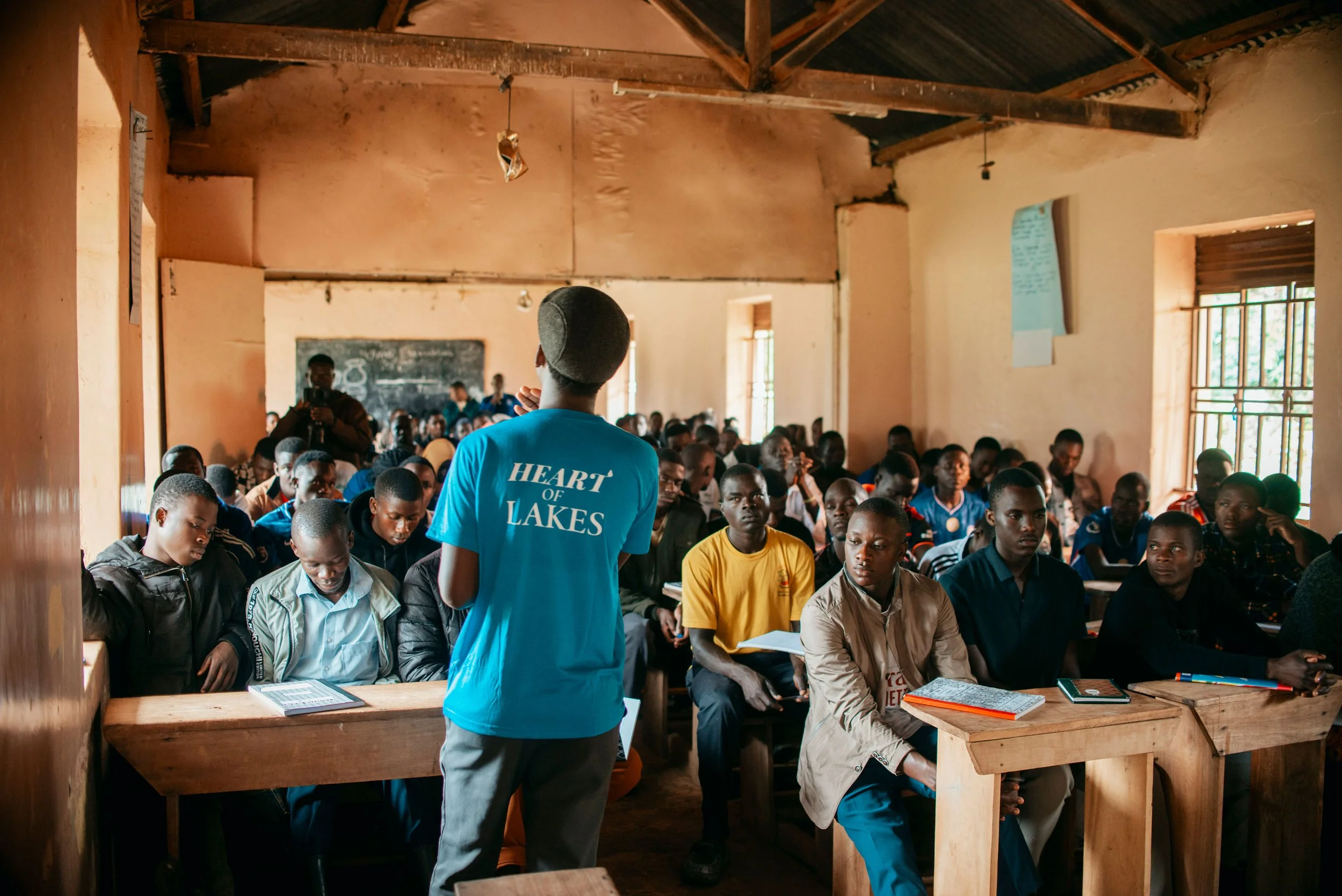 A classroom filled with students sitting at desks and paying attention. A man in a bright blue shirt with 'Heart of Lakes' on the back is standing at the front, possibly the teacher or speaker. The classroom has wooden walls and a blackboard at the front. Several students are looking towards the front, some writing, and others listening.
