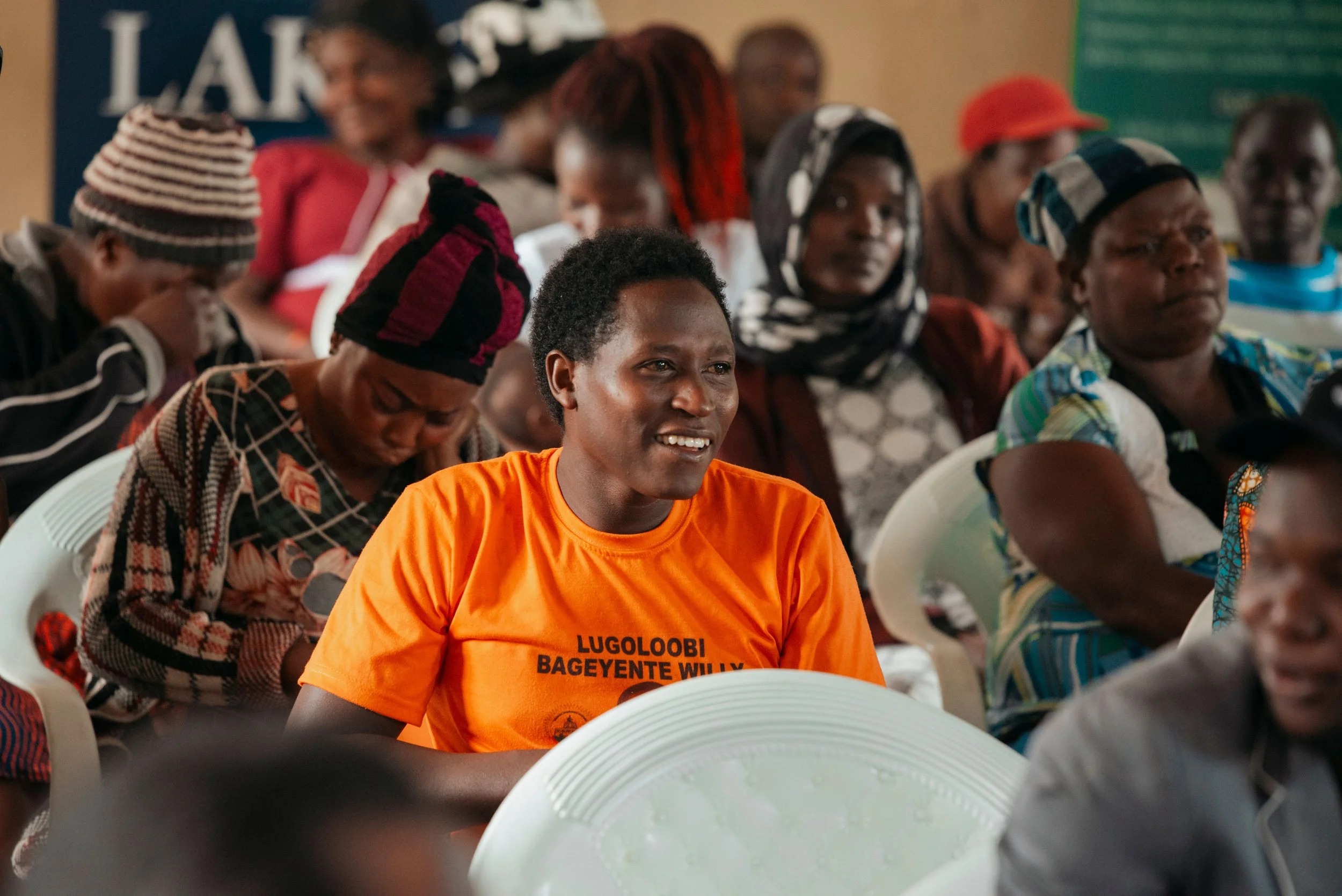 A group of African women and girls gathered indoors, seated on white chairs, some wearing colorful head coverings. One woman in the forefront is smiling and wearing an orange T-shirt with black text. The background includes a banner and a green bulletin board.