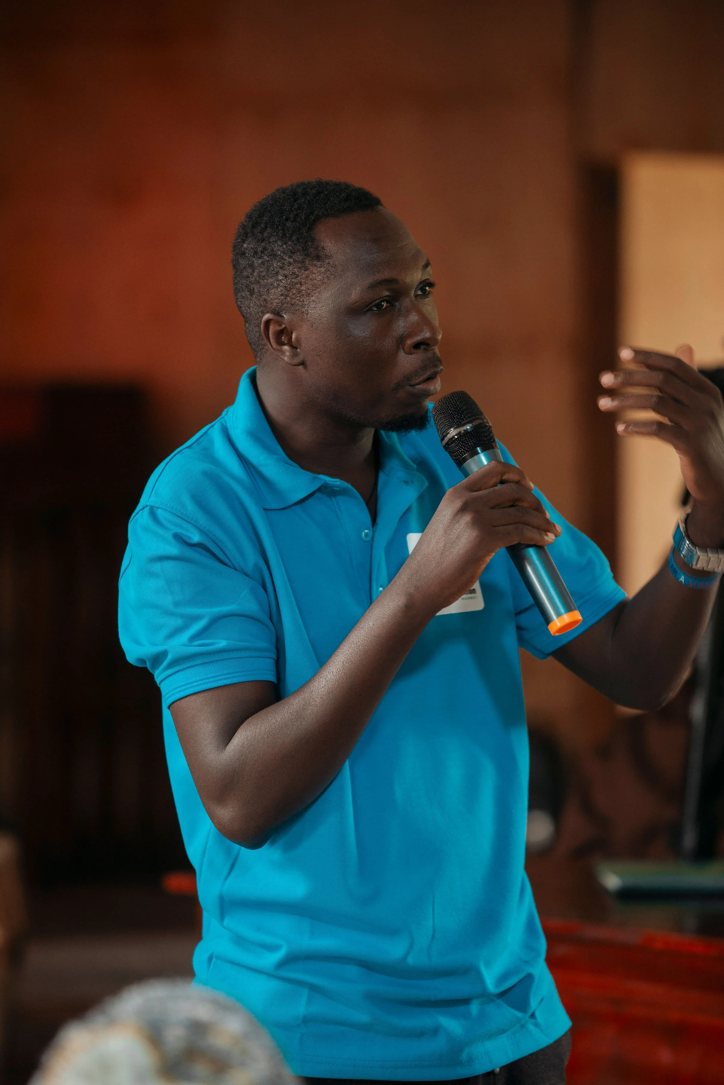 A man with short black hair and dark skin wearing a bright blue polo shirt, holding a microphone in his right hand, speaking and gesturing with his left hand during a presentation or discussion.