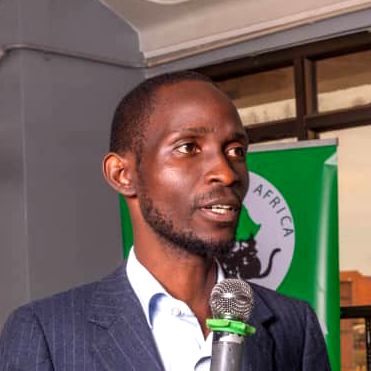 A man speaking into a microphone at an indoor event with an Africa-related banner in the background.