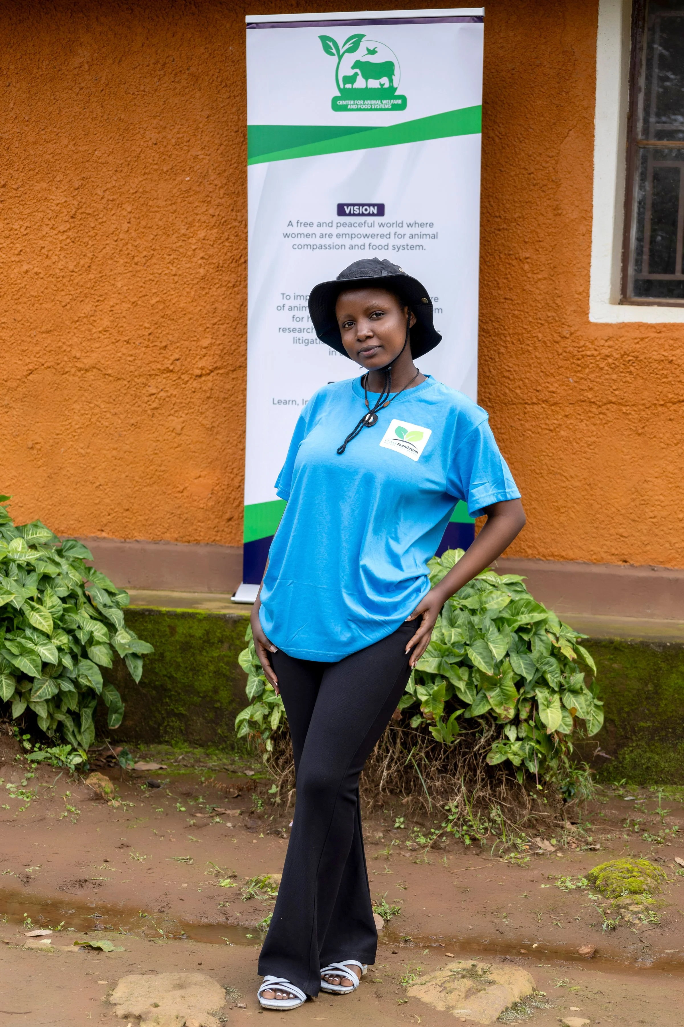 A woman in a blue shirt and black pants standing outdoors in front of an orange wall and a banner about animal welfare. She is wearing a black hat and white sandals.