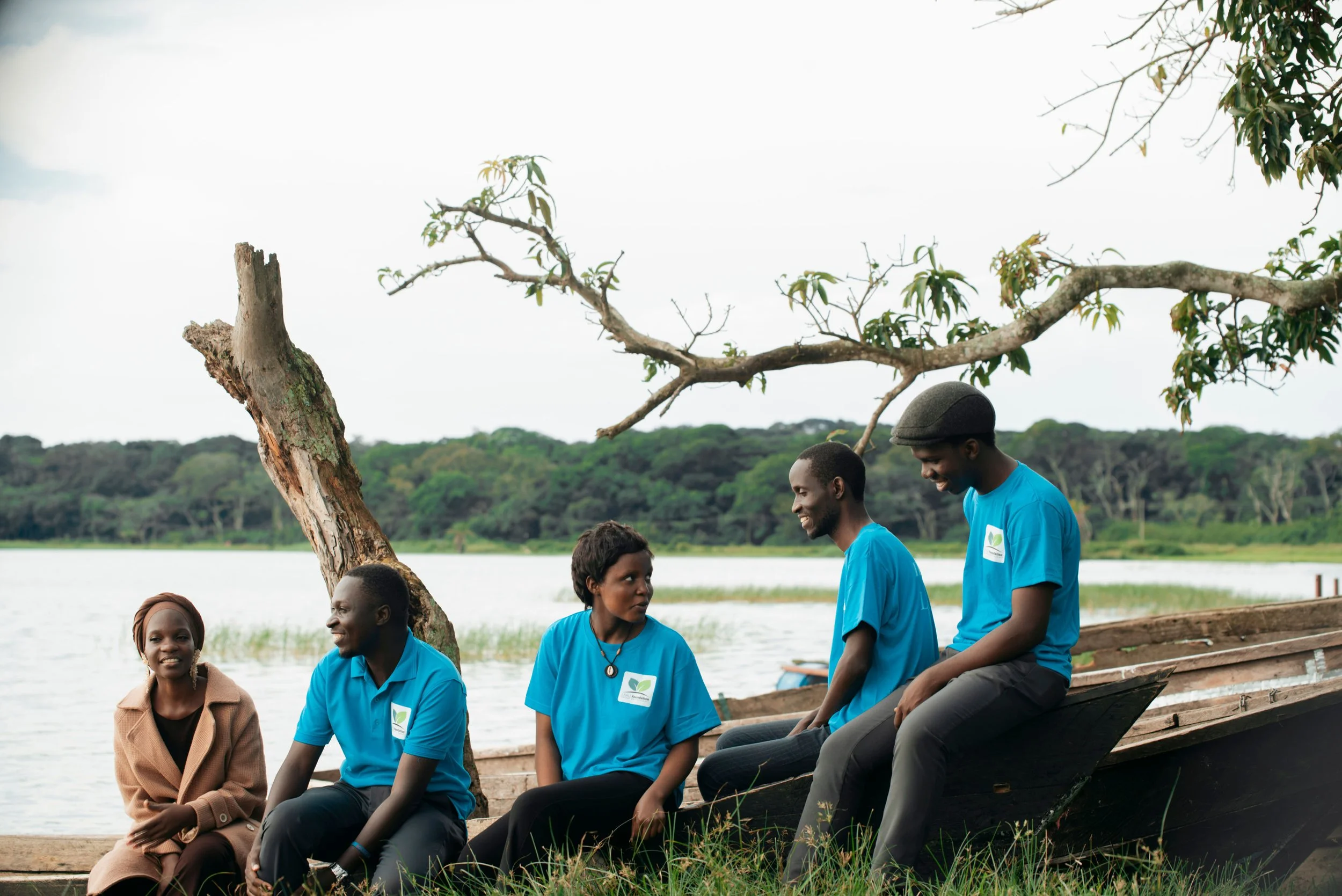 A group of six people sitting on a boat by a river, engaging in conversation, with trees and a distant riverbank in the background.
