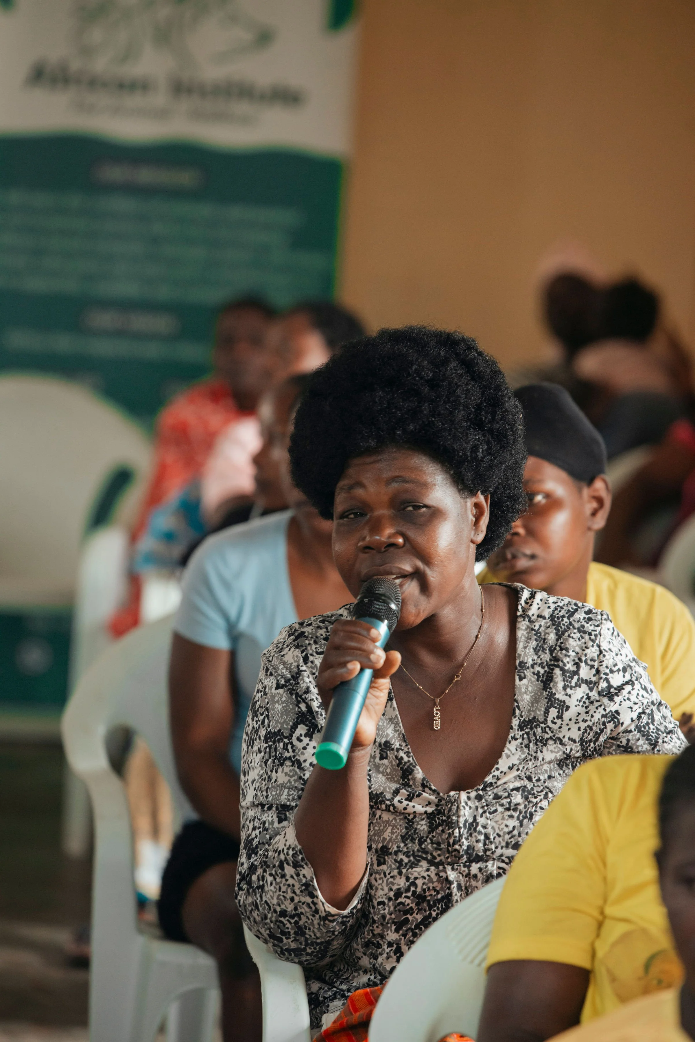 A woman speaking into a microphone at a meeting, with several people sitting in chairs behind her.
