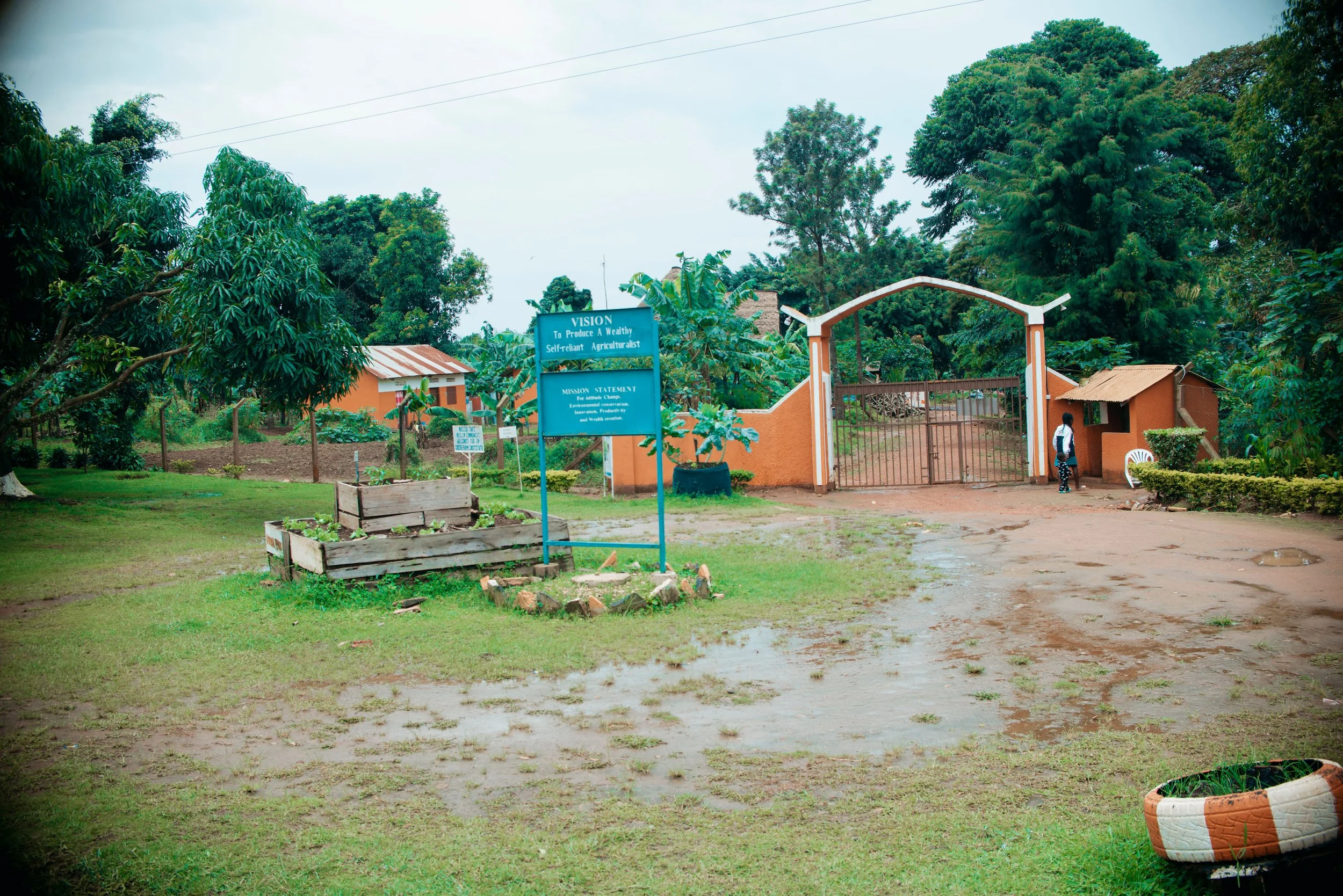 A rural scene with trees, a small building, and a gate with a person standing near it. There is a blue sign with mission statement and a garden bed in the foreground.