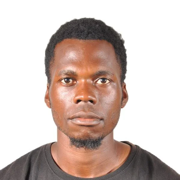 A young man with short curly hair and a goatee, wearing a black t-shirt, looking directly at the camera with a neutral expression, against a white background.