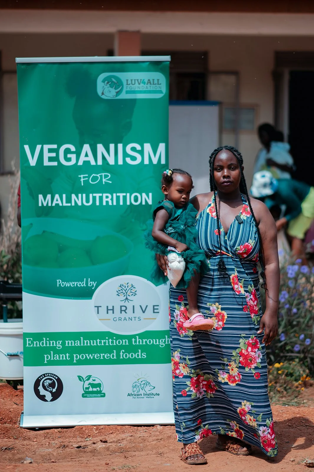 A woman standing outdoors holding a young girl in front of a green banner that reads "Veganism for Malnutrition," with logos and text promoting plant-powered foods to end malnutrition. The woman wears a floral dress, and the girl wears a dark green dress. Other people are visible in the background.