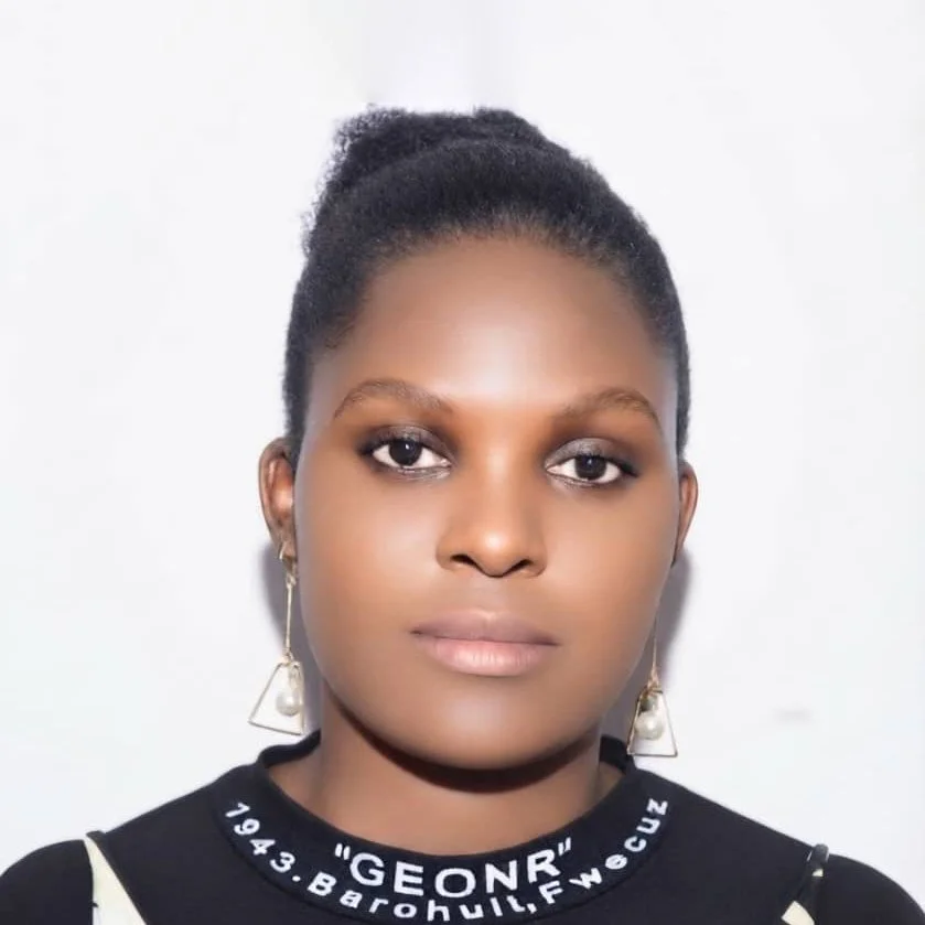A young woman with dark skin and natural hair tied back, wearing earrings and a black shirt with white lettering, standing against a plain white background.