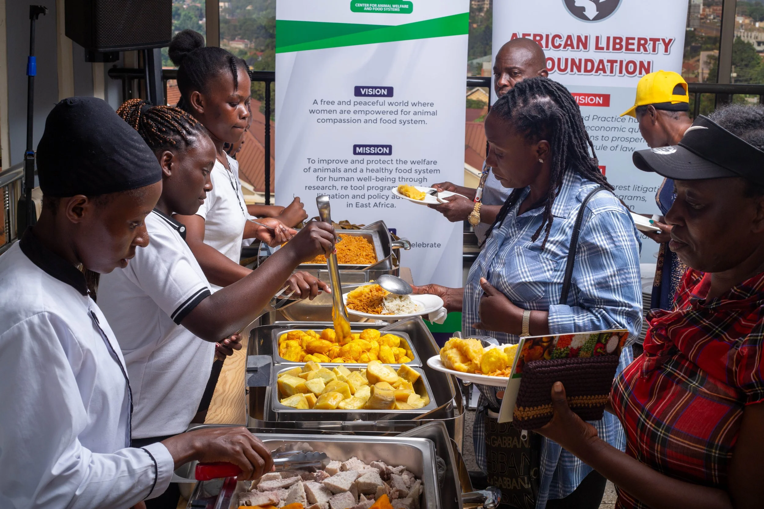 People in a queue at a buffet-style food service, with buffet trays filled with various foods and banners in the background.
